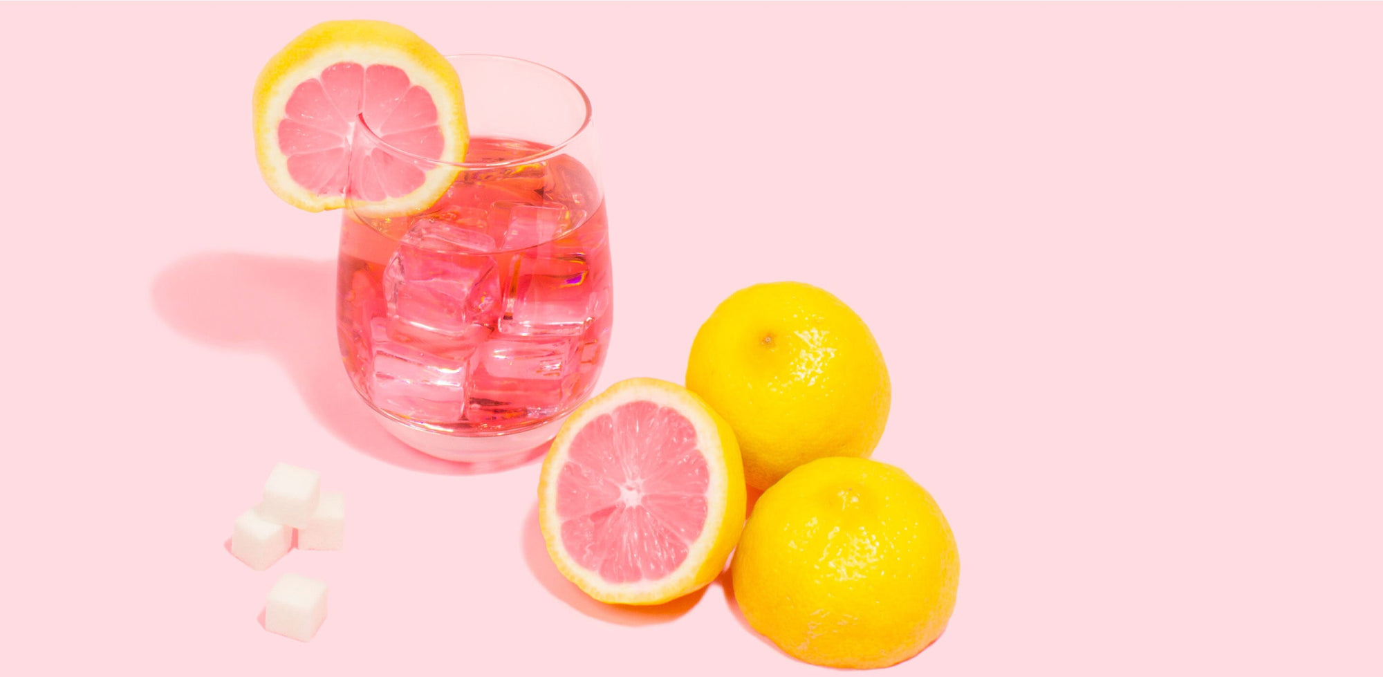 Glass of pink lemonade with ice cubes and lemon slices, surrounded by whole lemons and sugar cubes on a pastel pink background.