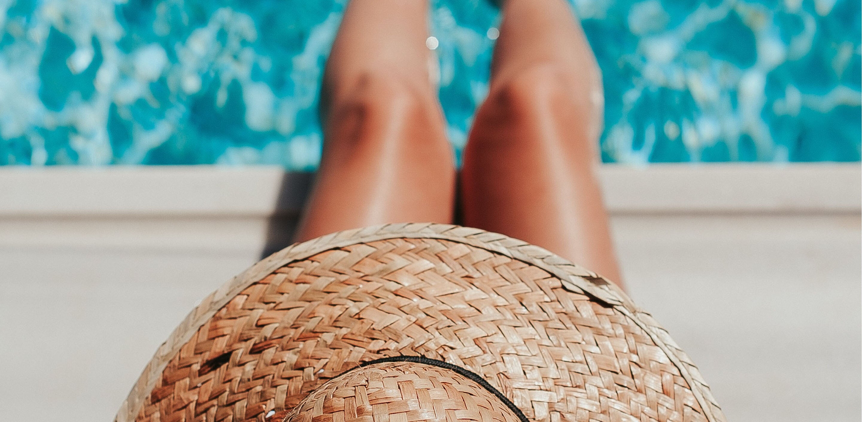 Person lounging poolside with a woven sun hat on their lap and legs stretched toward the water.