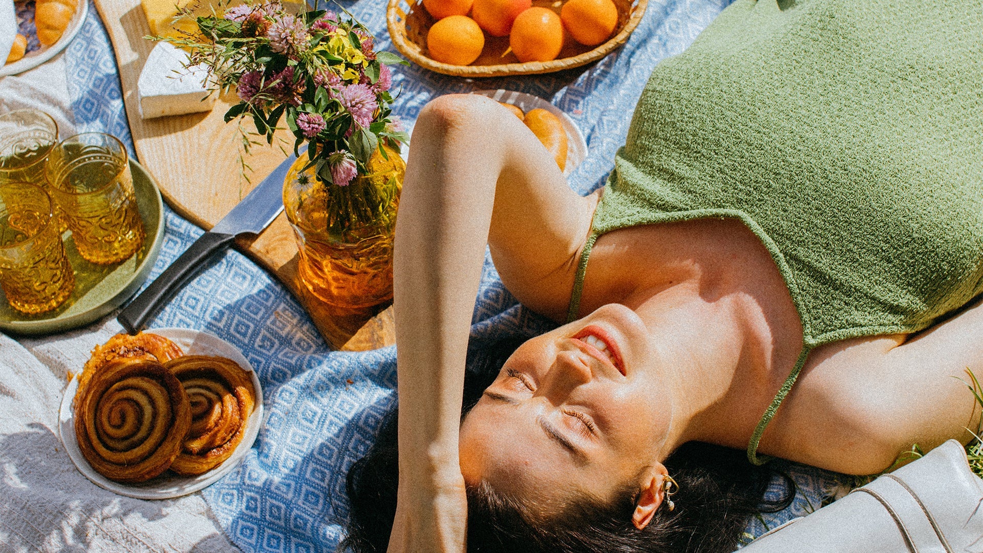 A woman lies on her back on a blanket with sun on her face, next to cinnamon rolls and oranges