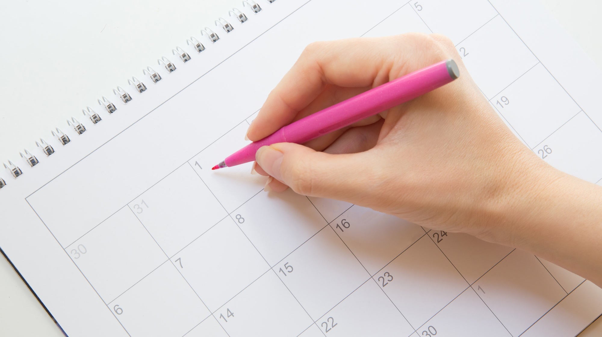 Hand holding a pink pen and pointing to a date on a blank calendar page.