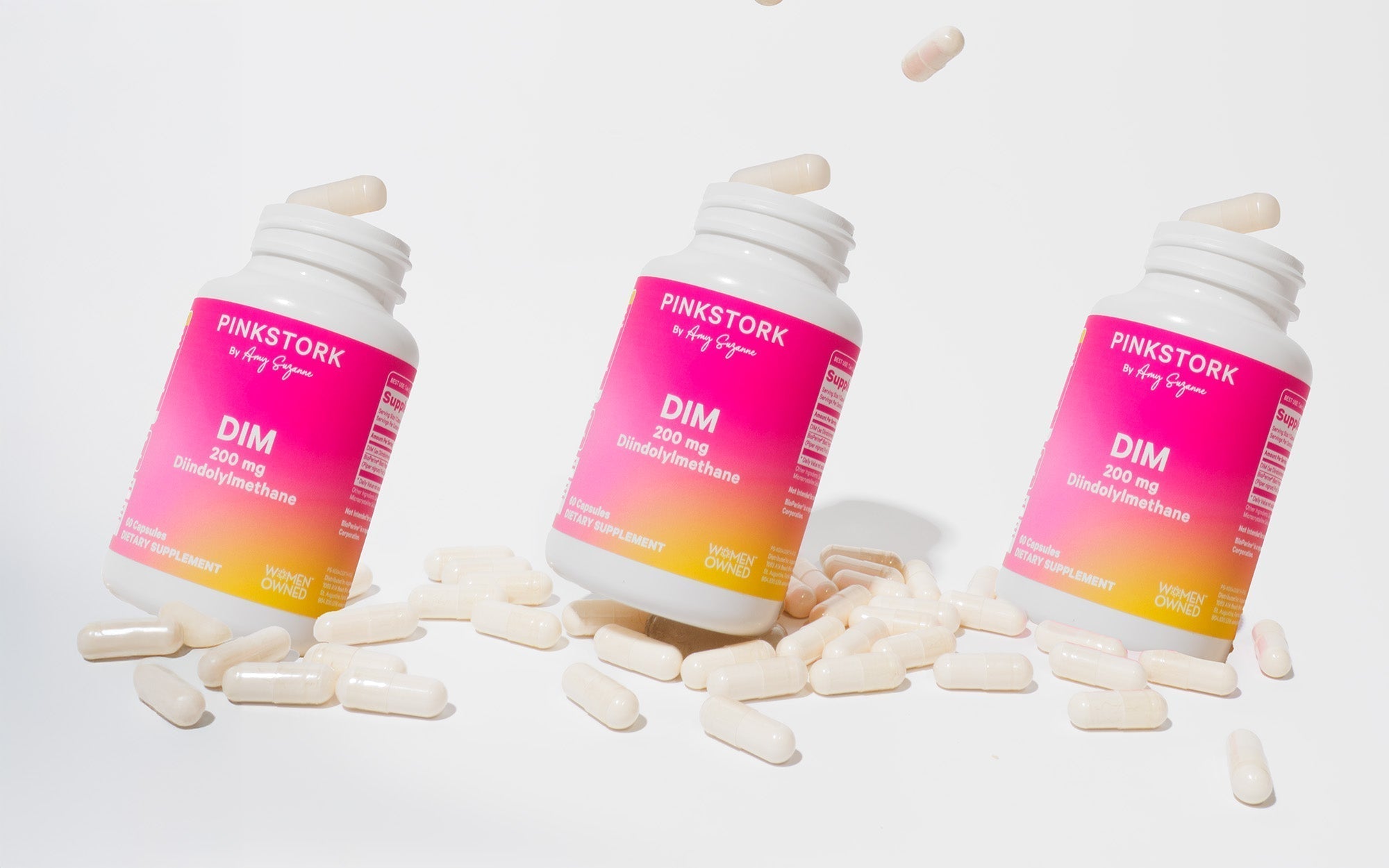 Three bottles of Pink Stork DIM 200 mg dietary supplement with white capsules scattered on a white background.