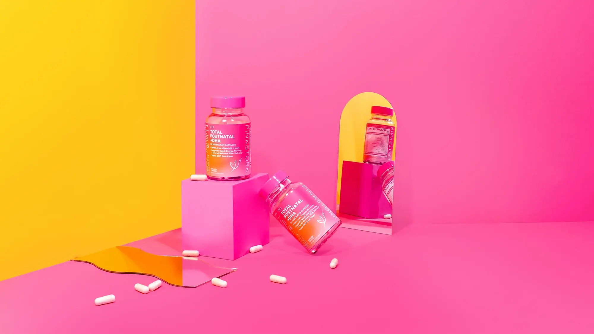 Pink Stork Total Prenatal vitamin supplement bottles with capsules on colorful pink and yellow background with mirror reflection.