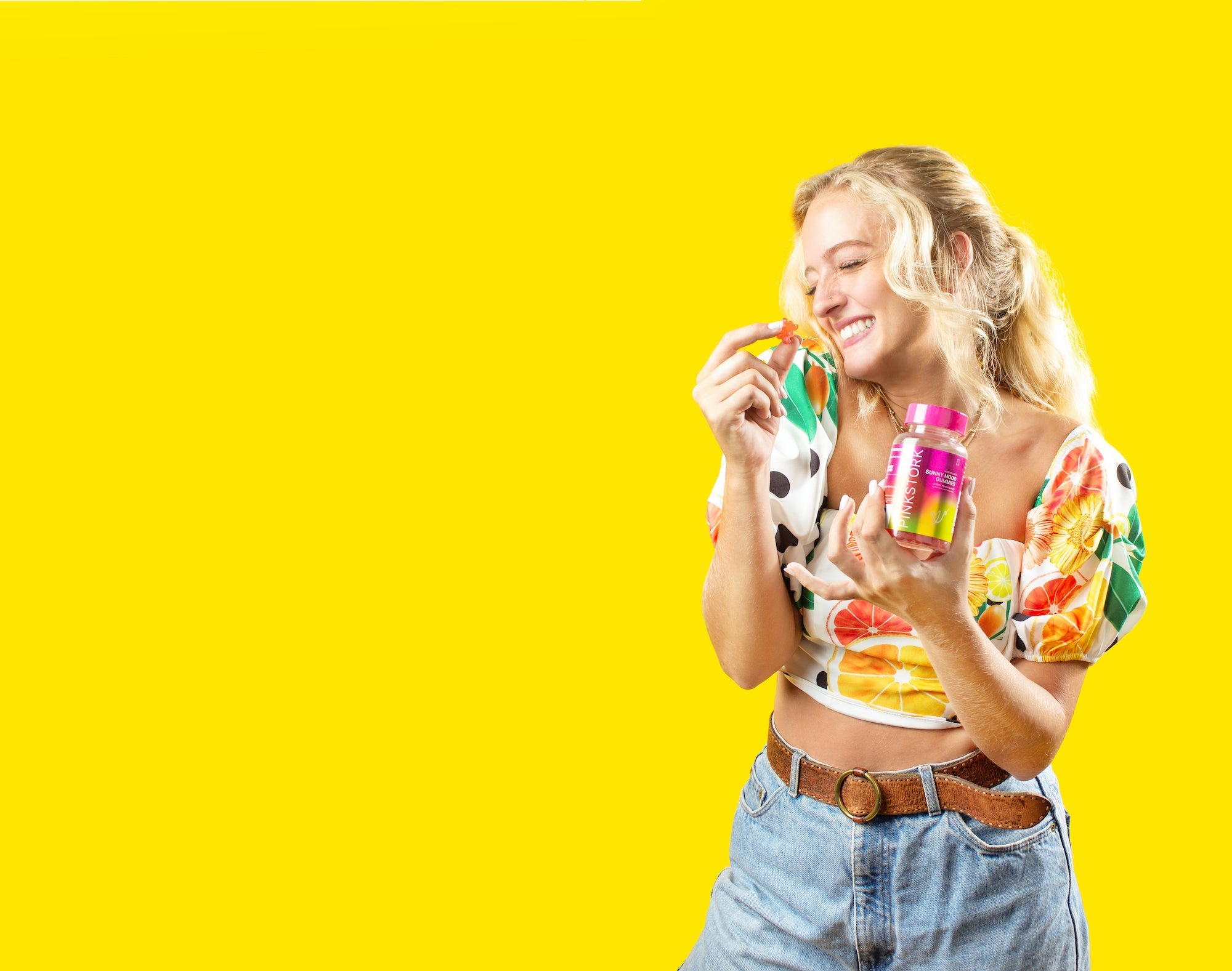 Smiling woman in a floral crop top holding a Pink Stork supplement bottle and gummy against a bright yellow background.