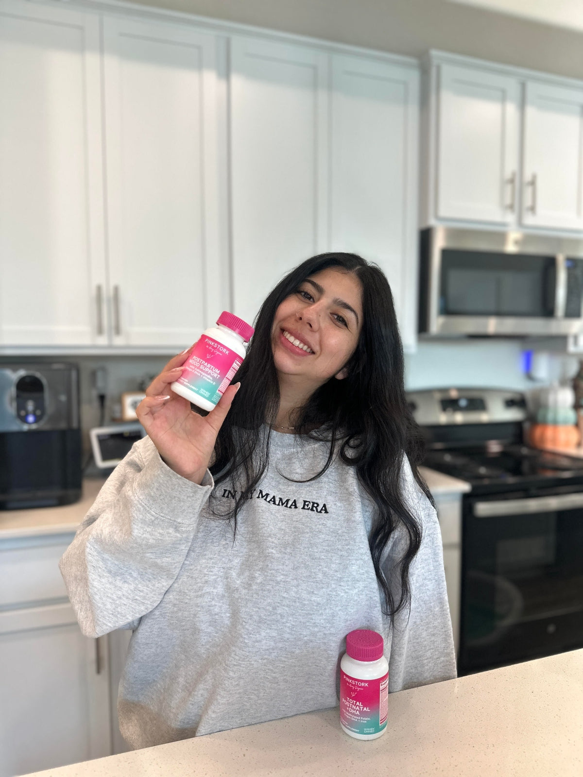Smiling woman in a gray ‘In My Mama Era’ sweatshirt holding a Pink Stork supplement bottle in a modern kitchen.