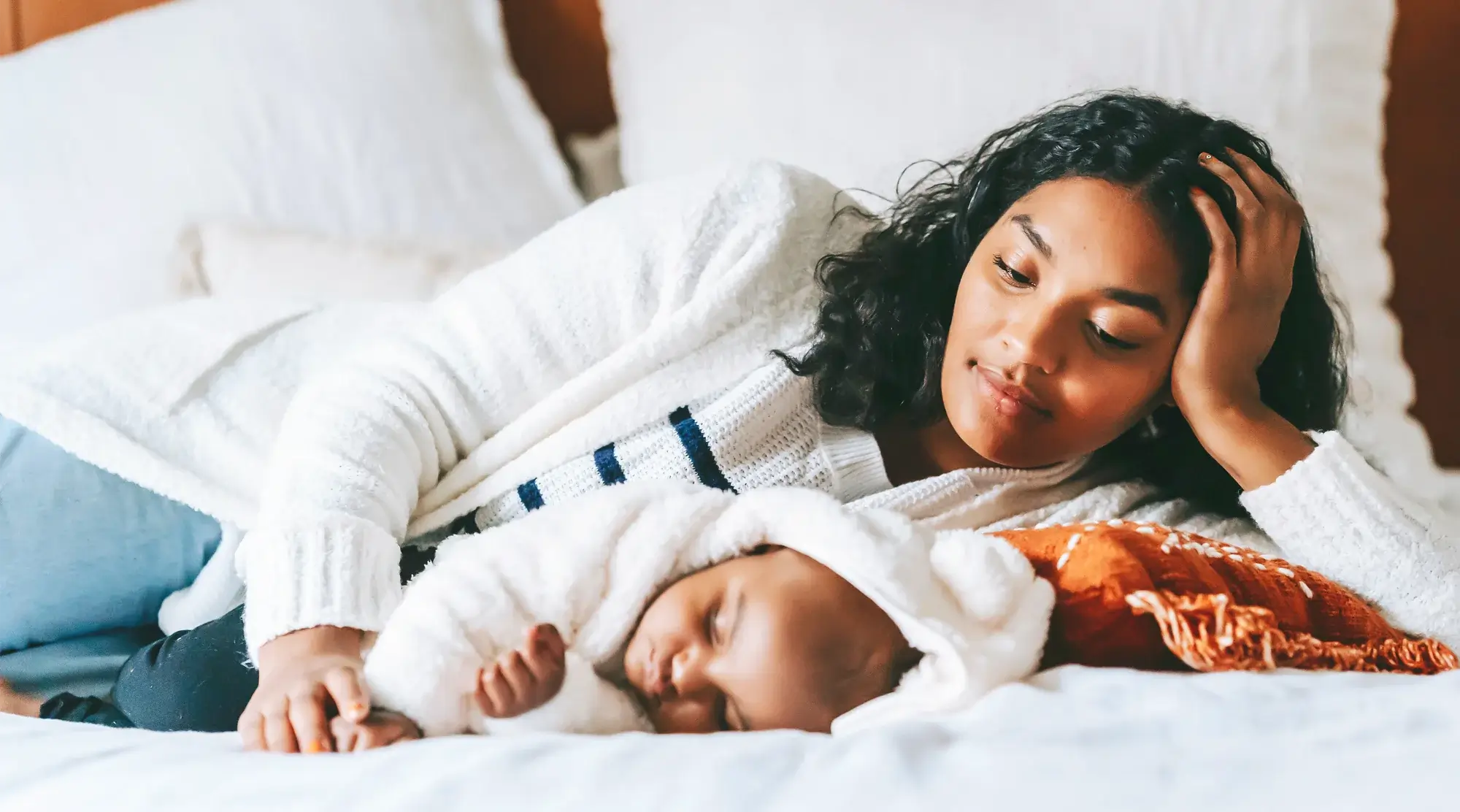 Mother resting on bed while watching her sleeping baby in cozy white and orange bedding.