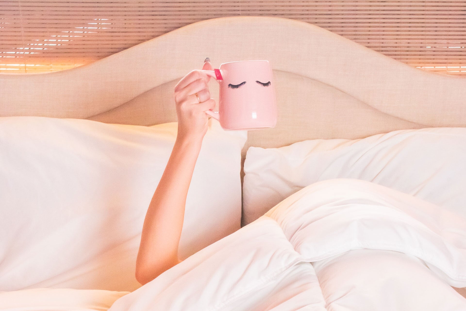 Person lying under white bedding holding up a pink coffee mug with closed-eye design.
