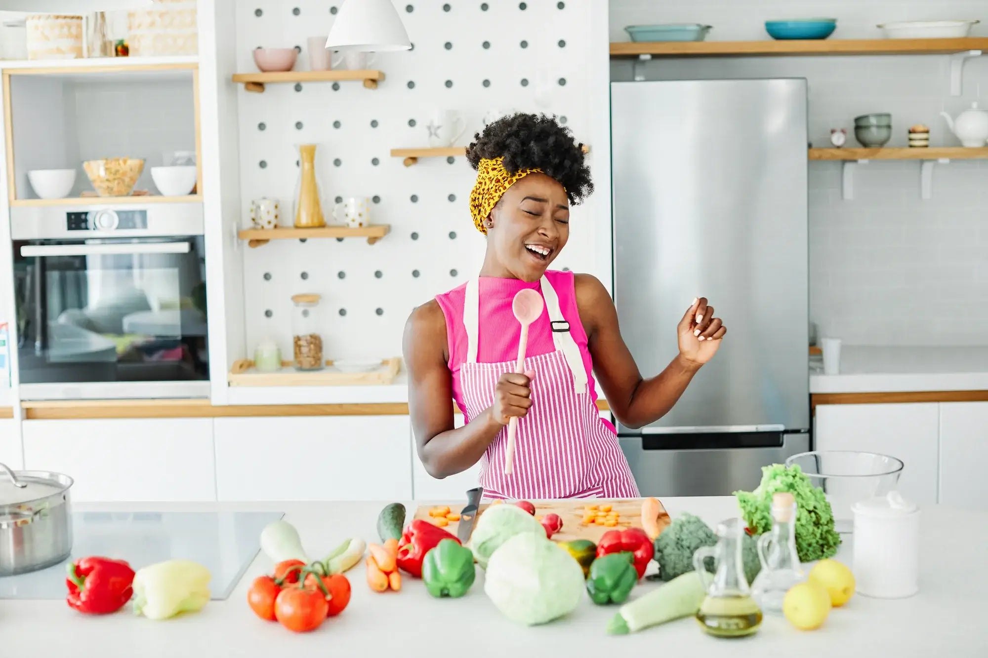 Joyful woman in pink apron singing while preparing fresh vegetables in modern kitchen.