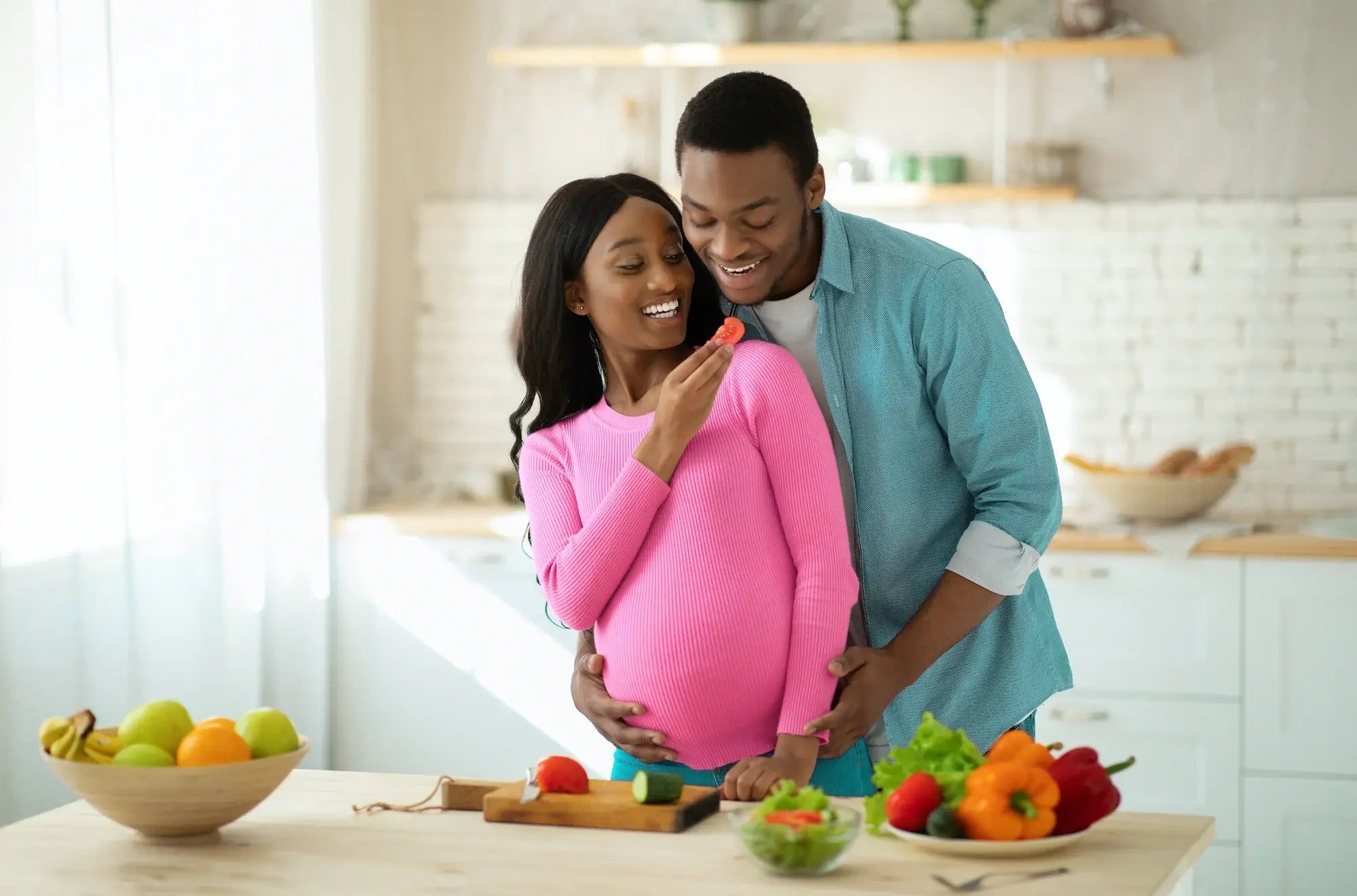 Happy pregnant couple preparing healthy meal together in kitchen surrounded by fresh vegetables and fruits.