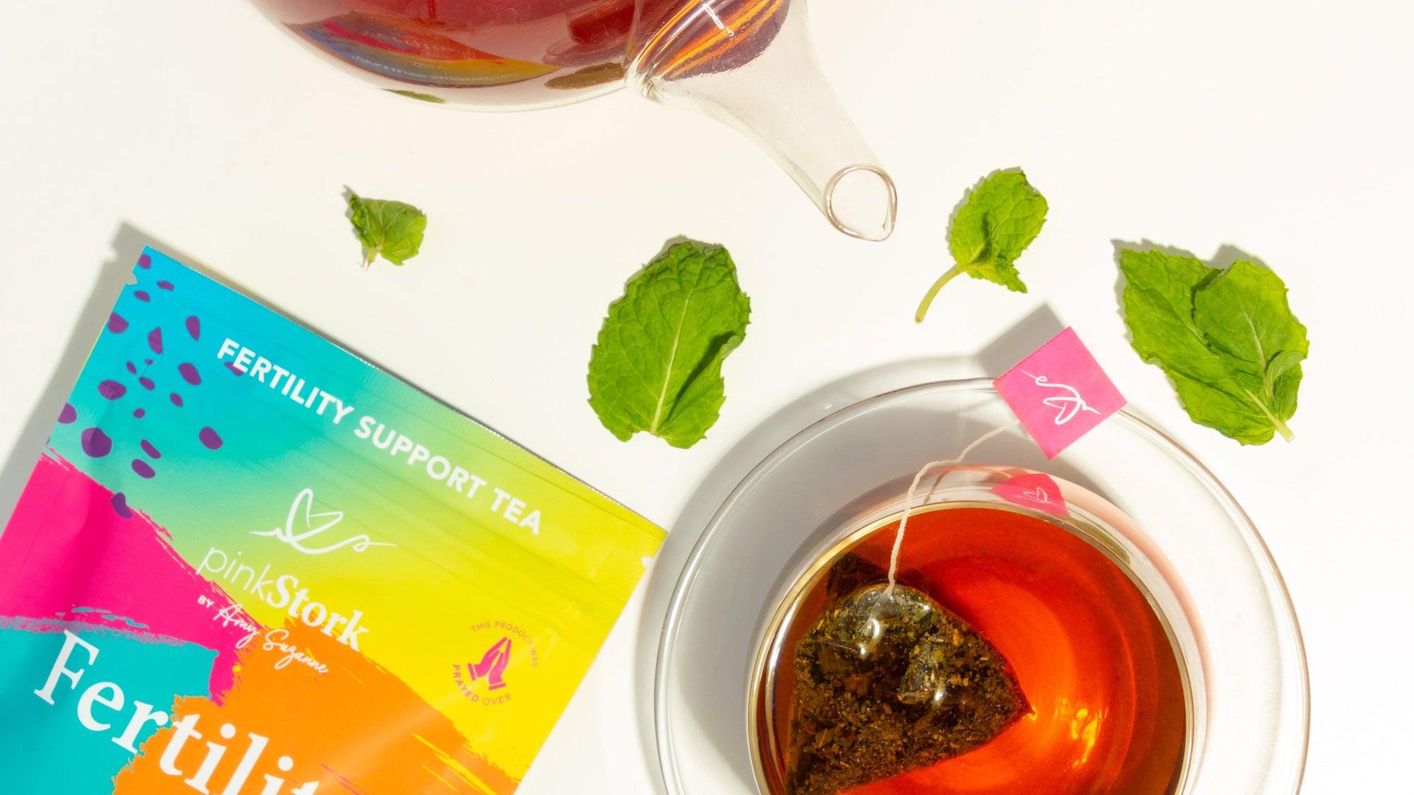 Pink Stork Fertility Support Tea with colorful packaging, a cup of brewed tea, teapot, and fresh mint leaves.