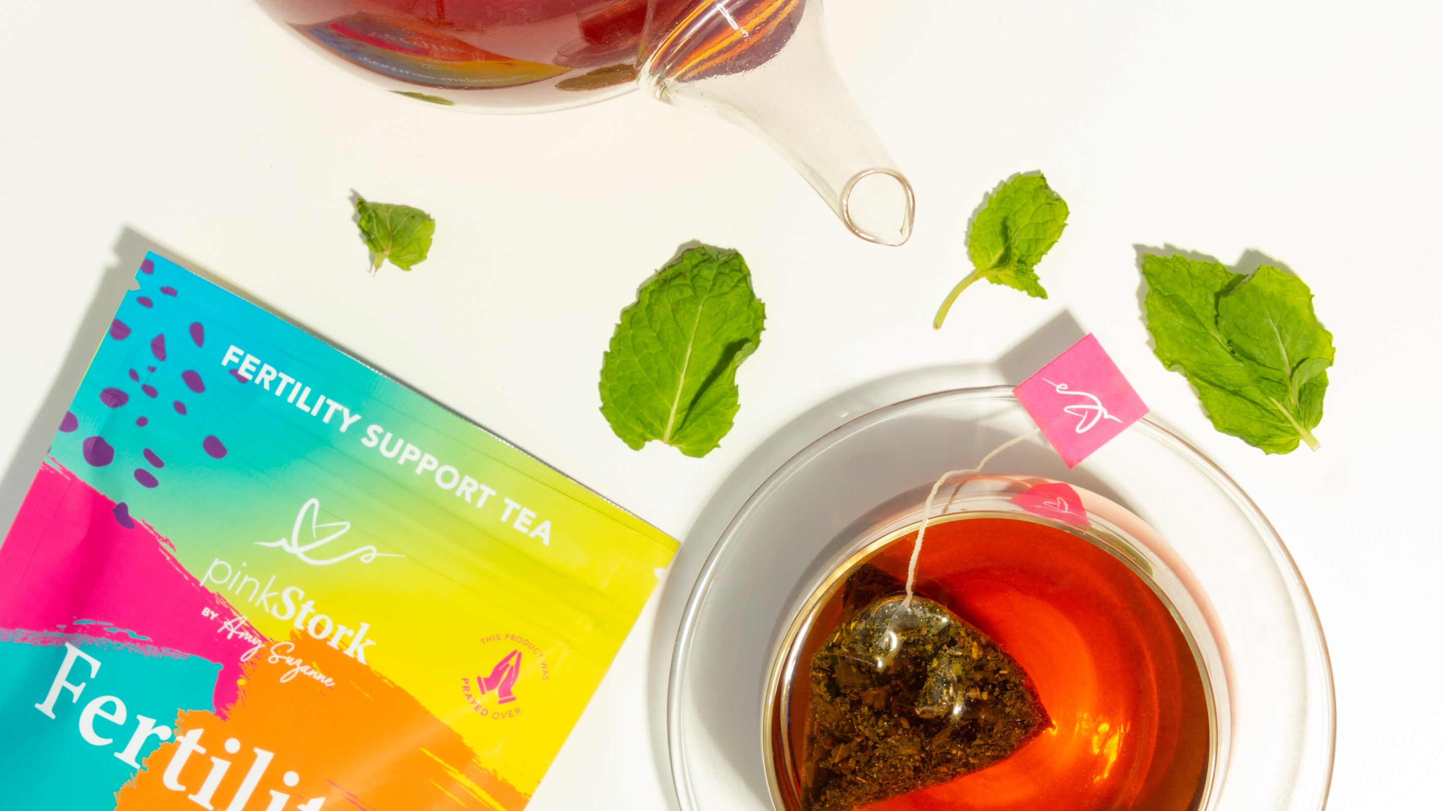Pink Stork Fertility Support Tea with colorful packaging, a cup of brewed tea, teapot, and fresh mint leaves.