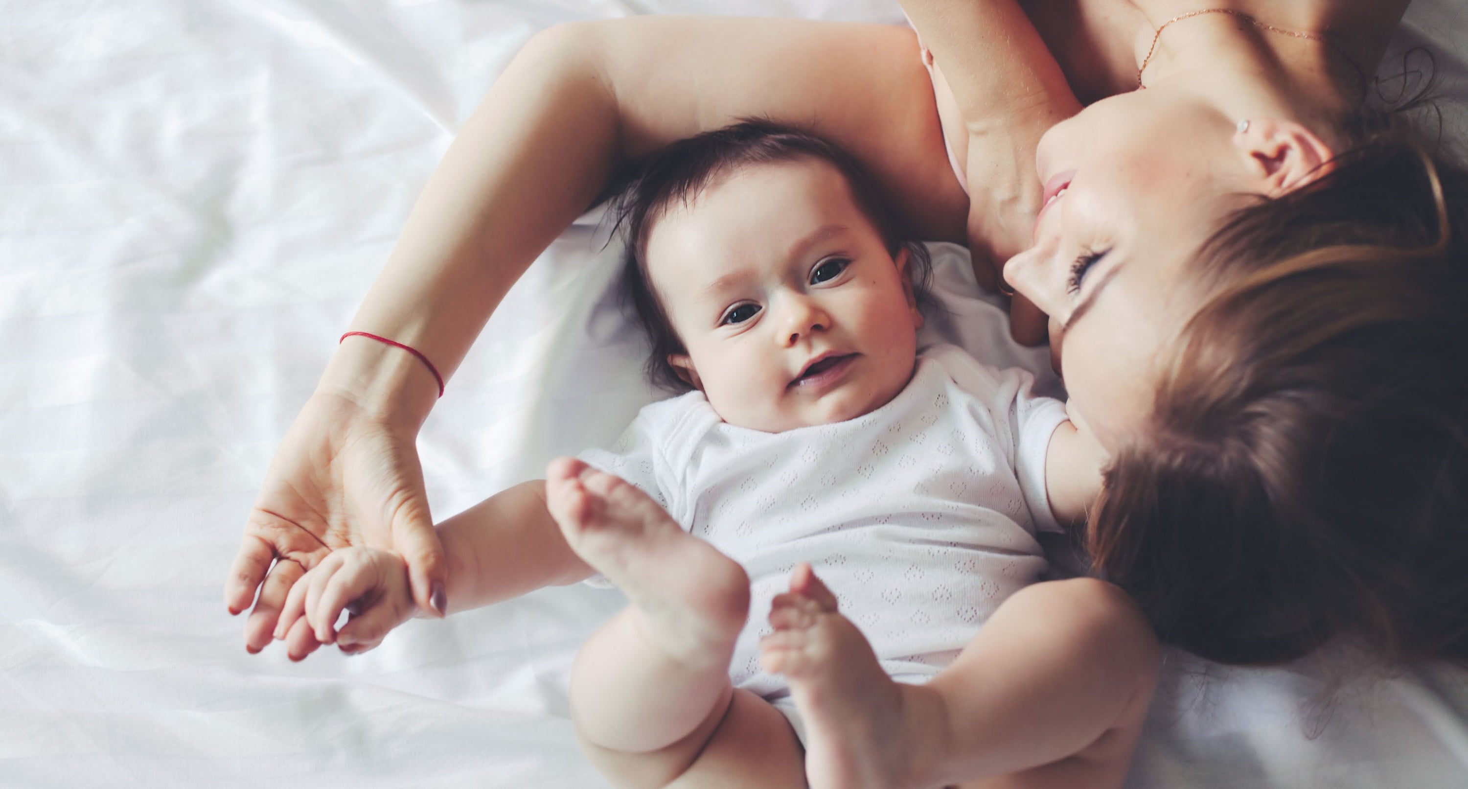 Mother lying on a bed next to her baby, both dressed in white, with the baby looking at the camera while the mother smiles affectionately at her child.