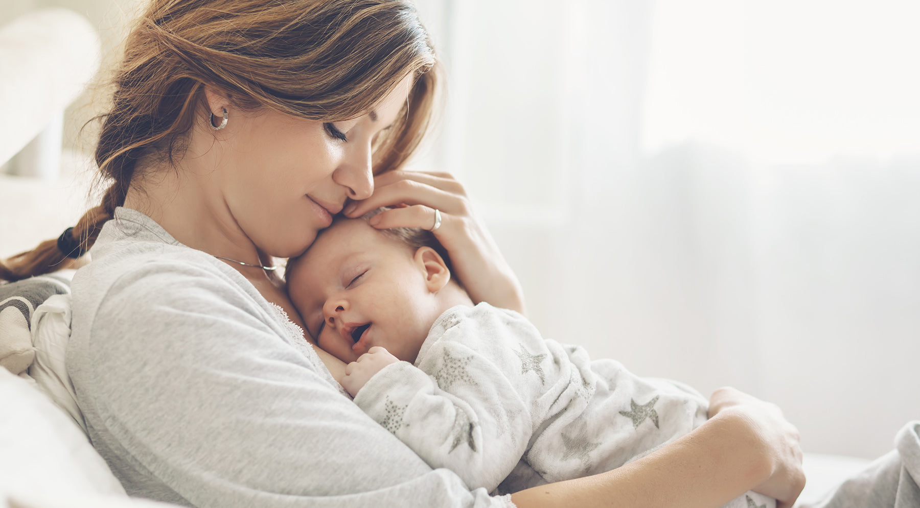 Mother cuddling her sleeping baby in cozy loungewear near a sunlit window.