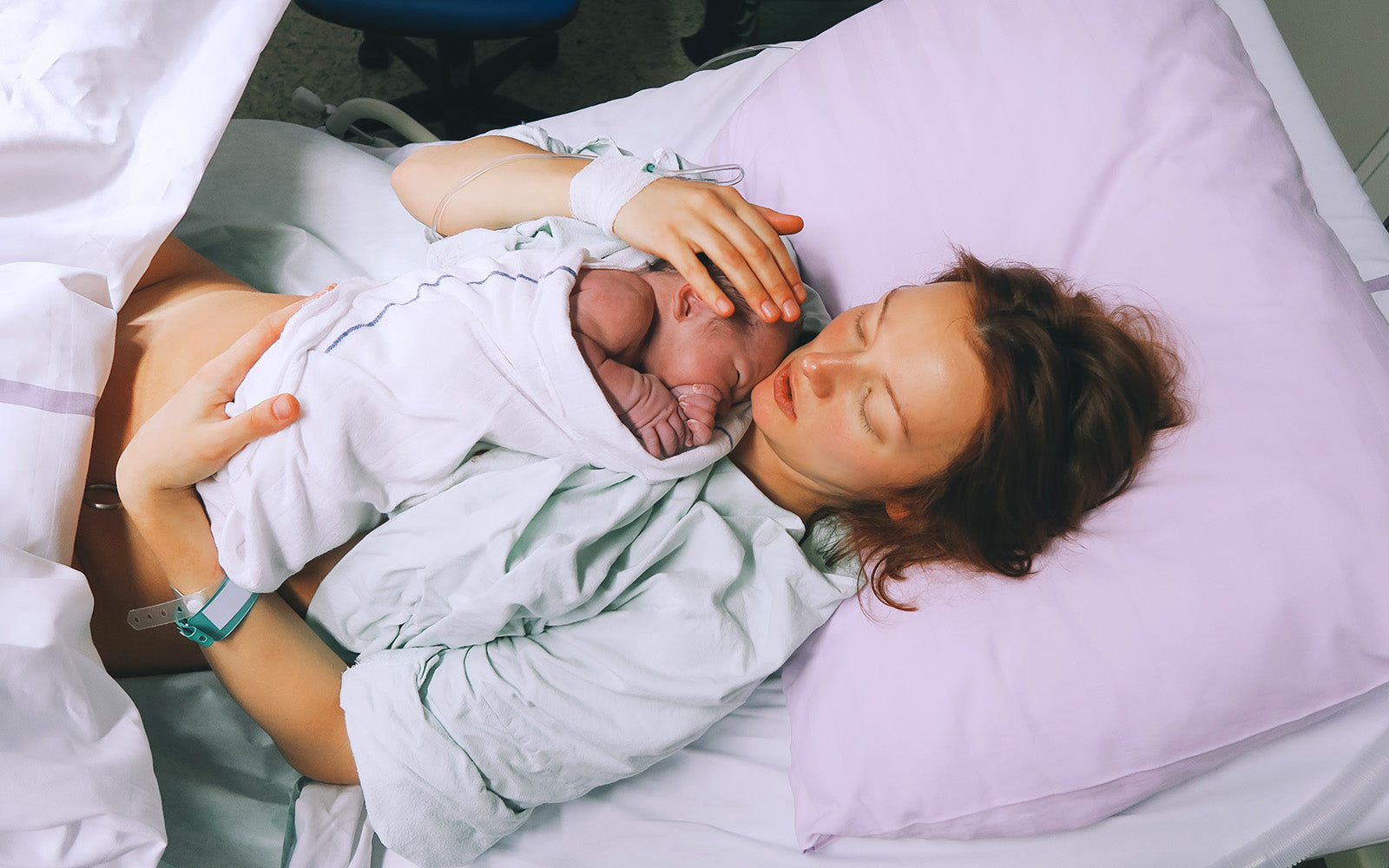 New mother resting in hospital bed while holding newborn baby wrapped in a blanket.