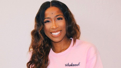 Smiling woman with long wavy hair wearing a pink Shakeout sweatshirt against a plain background.
