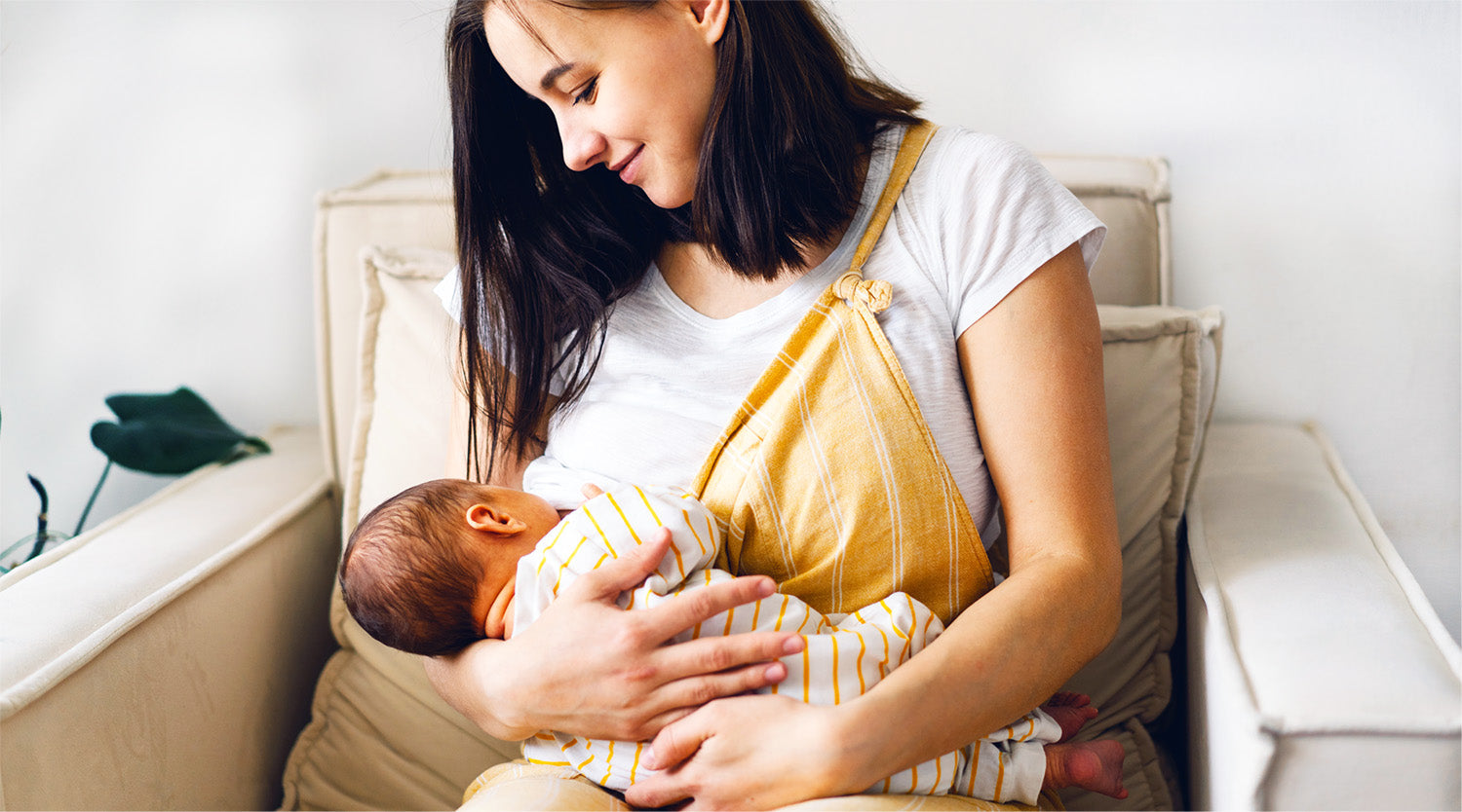 Smiling mother breastfeeding her newborn baby while sitting on a beige couch with a yellow sling wrap.