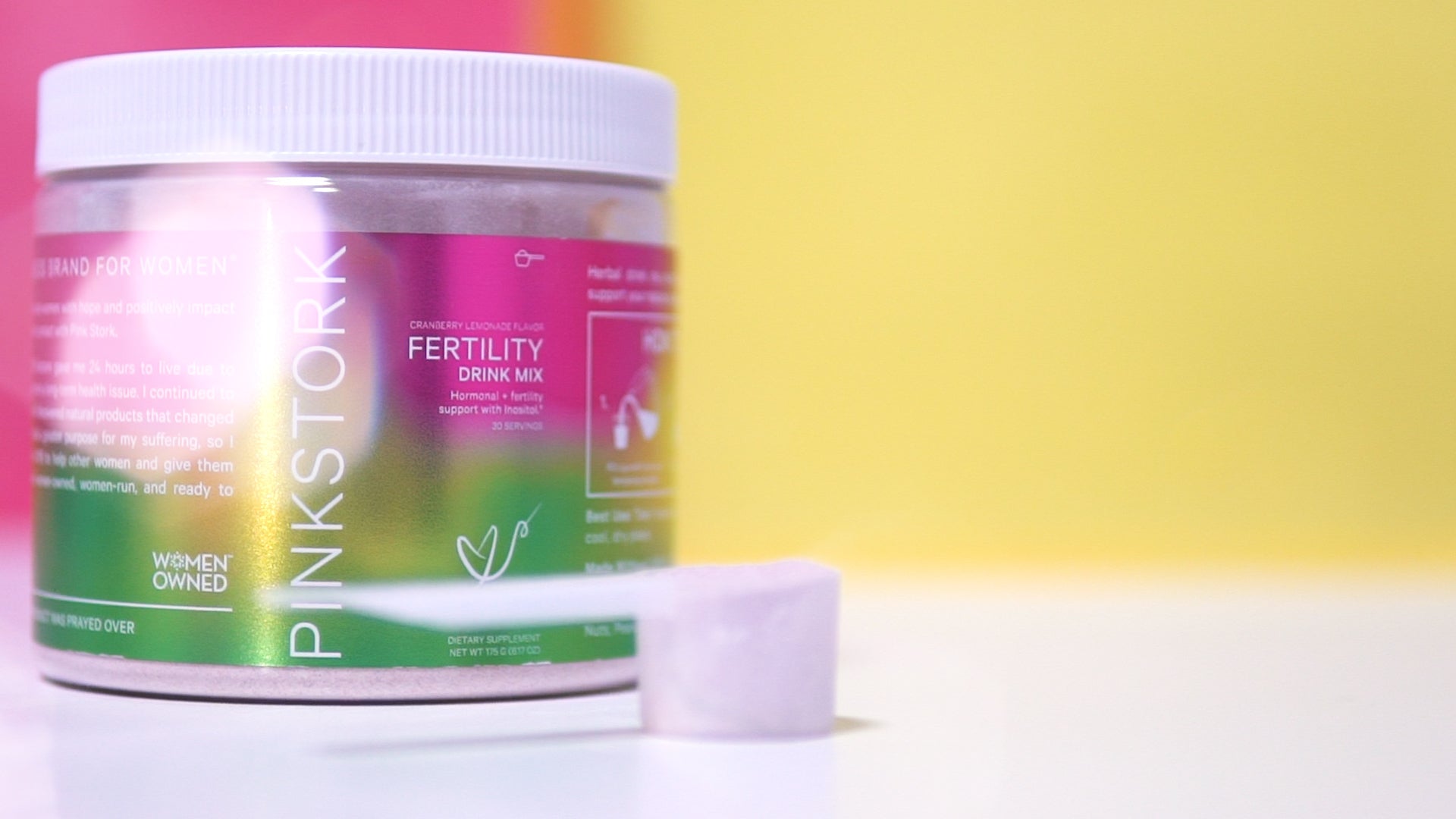 Pink Stork Fertility Drink Mix container with scoop on a white surface, colorful pink, green, and yellow background.