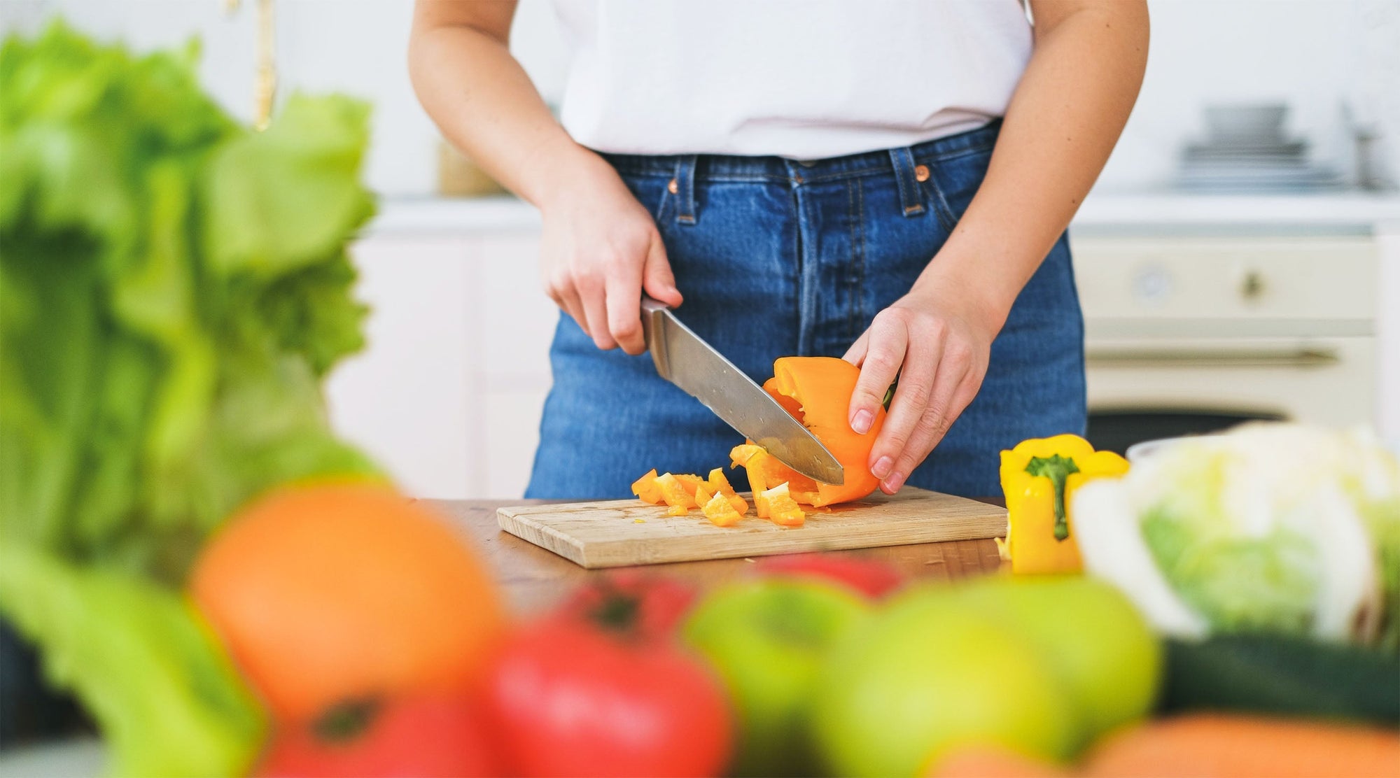 Person chopping fresh yellow bell pepper on a wooden cutting board surrounded by colorful vegetables in a kitchen.