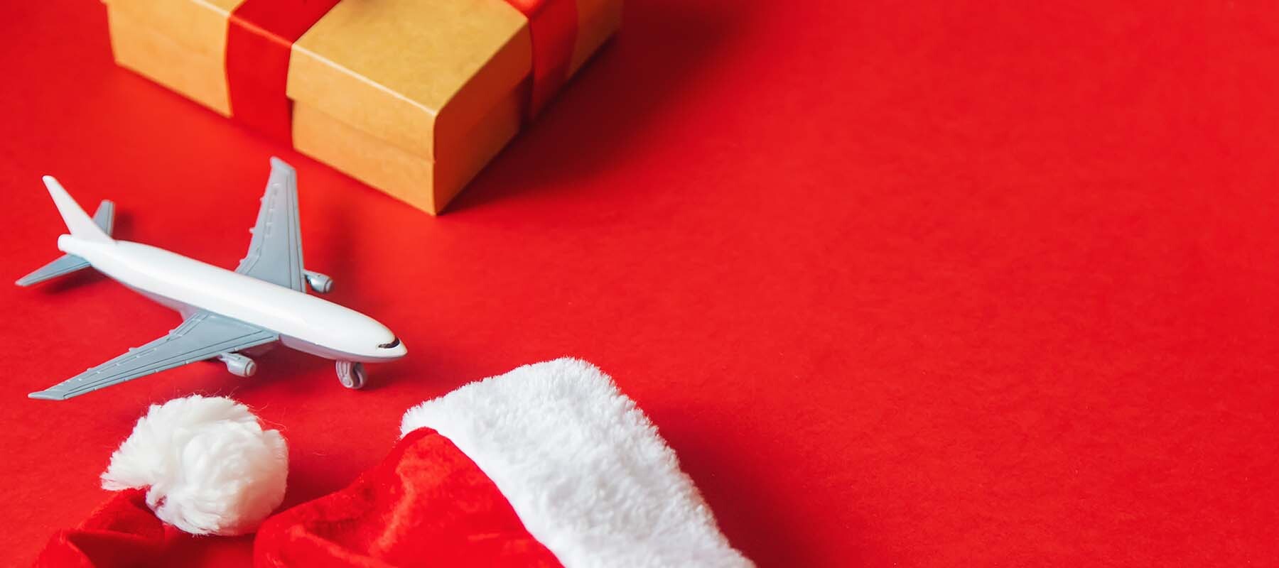 Miniature airplane, wrapped gift box, and Santa hat on a red background symbolizing Christmas holiday travel.