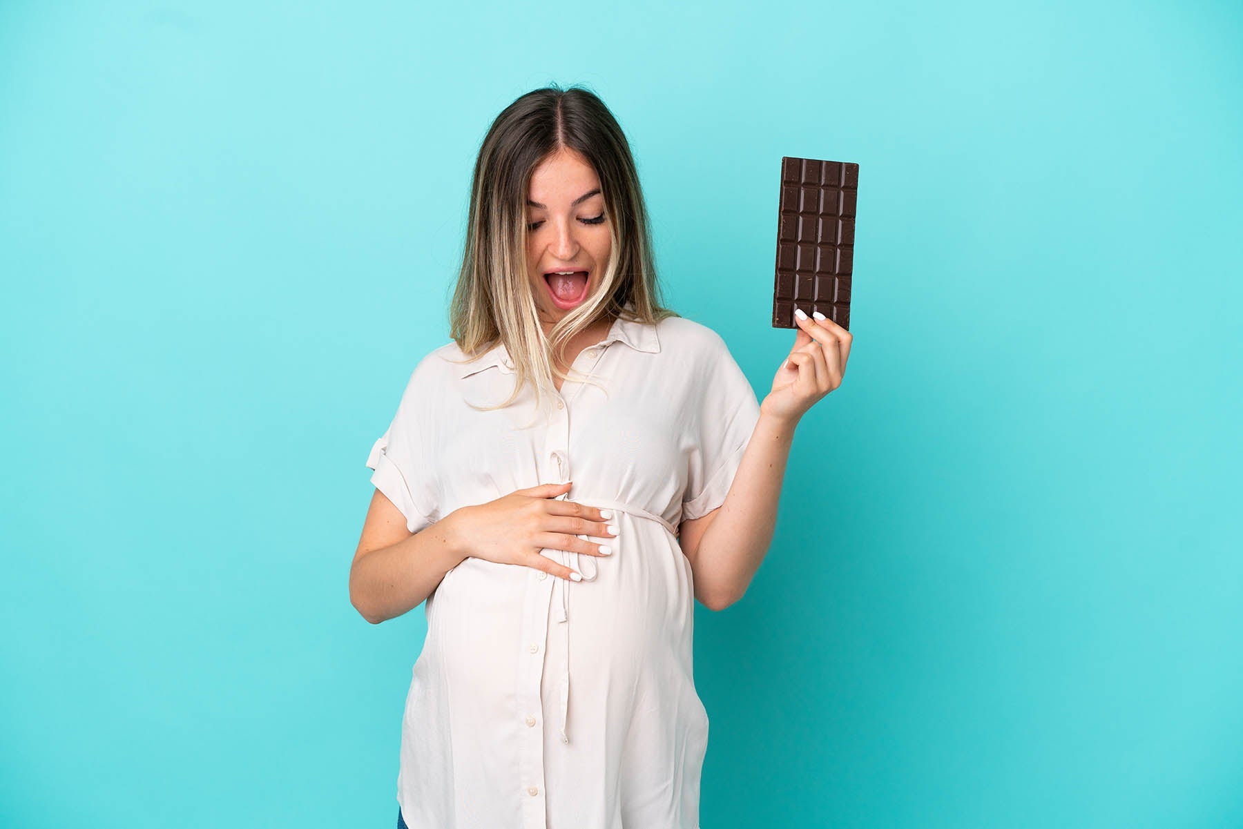 Excited pregnant woman holding a bar of chocolate against a blue background, representing pregnancy cravings and sweet indulgence.
