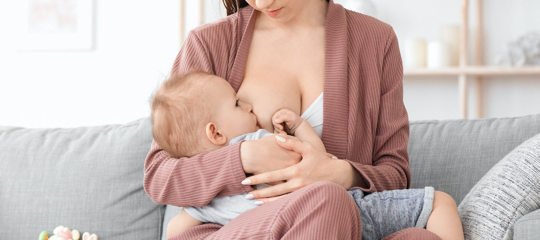 Mother in a mauve loungewear set breastfeeding her baby on a gray couch in a cozy home setting.