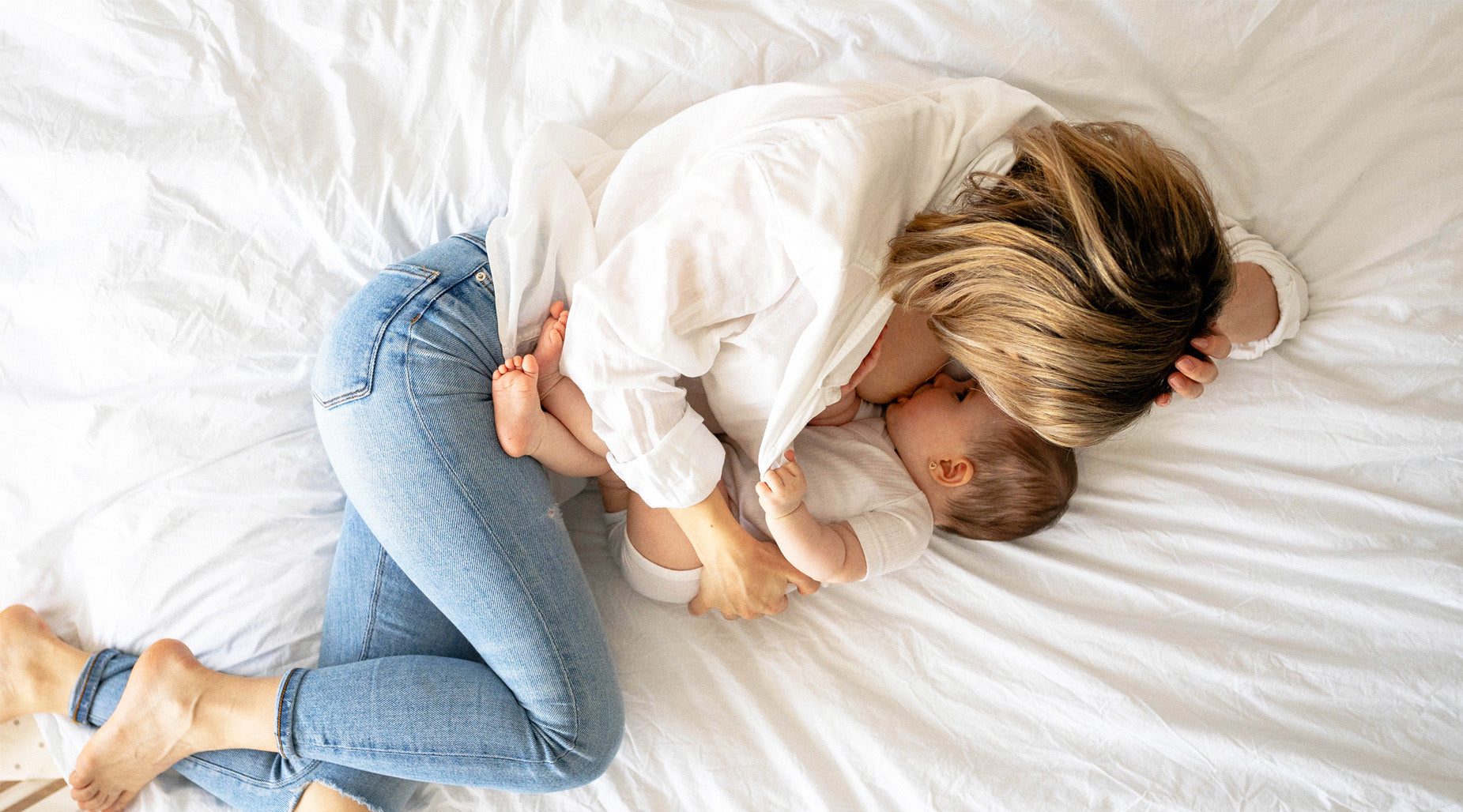 Mother lying on a bed breastfeeding her baby, dressed in a white shirt and jeans, sharing a nurturing skin-to-skin moment.