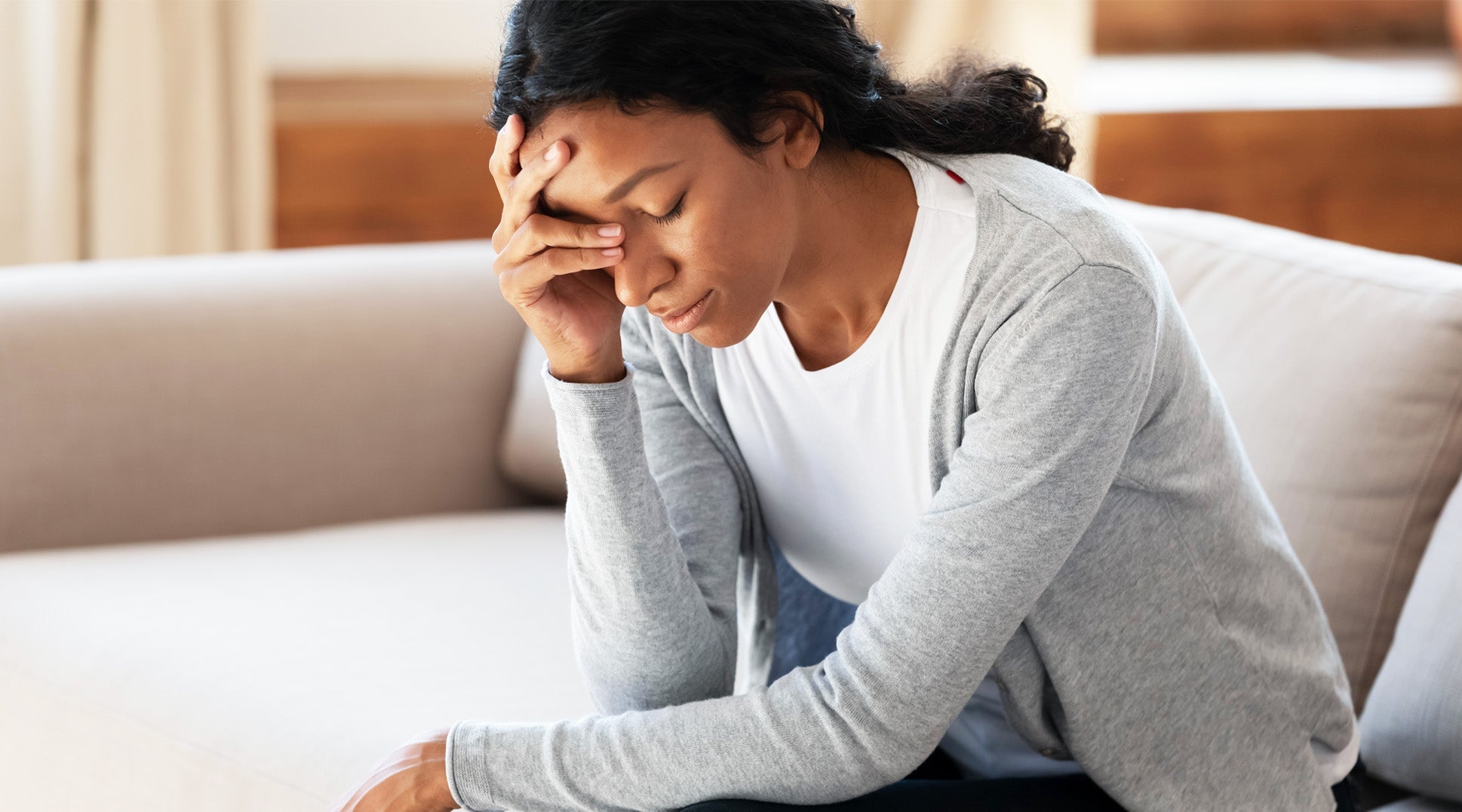 Stressed woman sitting on couch with hand on forehead, showing fatigue, anxiety, or mental exhaustion.