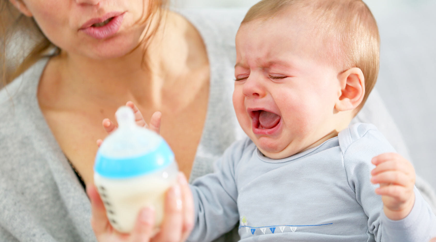 Crying baby in a blue onesie refusing a bottle of milk held by an adult.