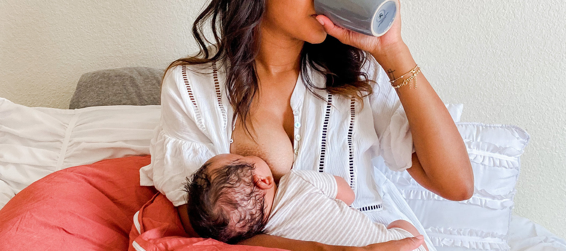 Mother breastfeeding her baby while drinking from a cup, sitting on a bed with coral and white pillows.