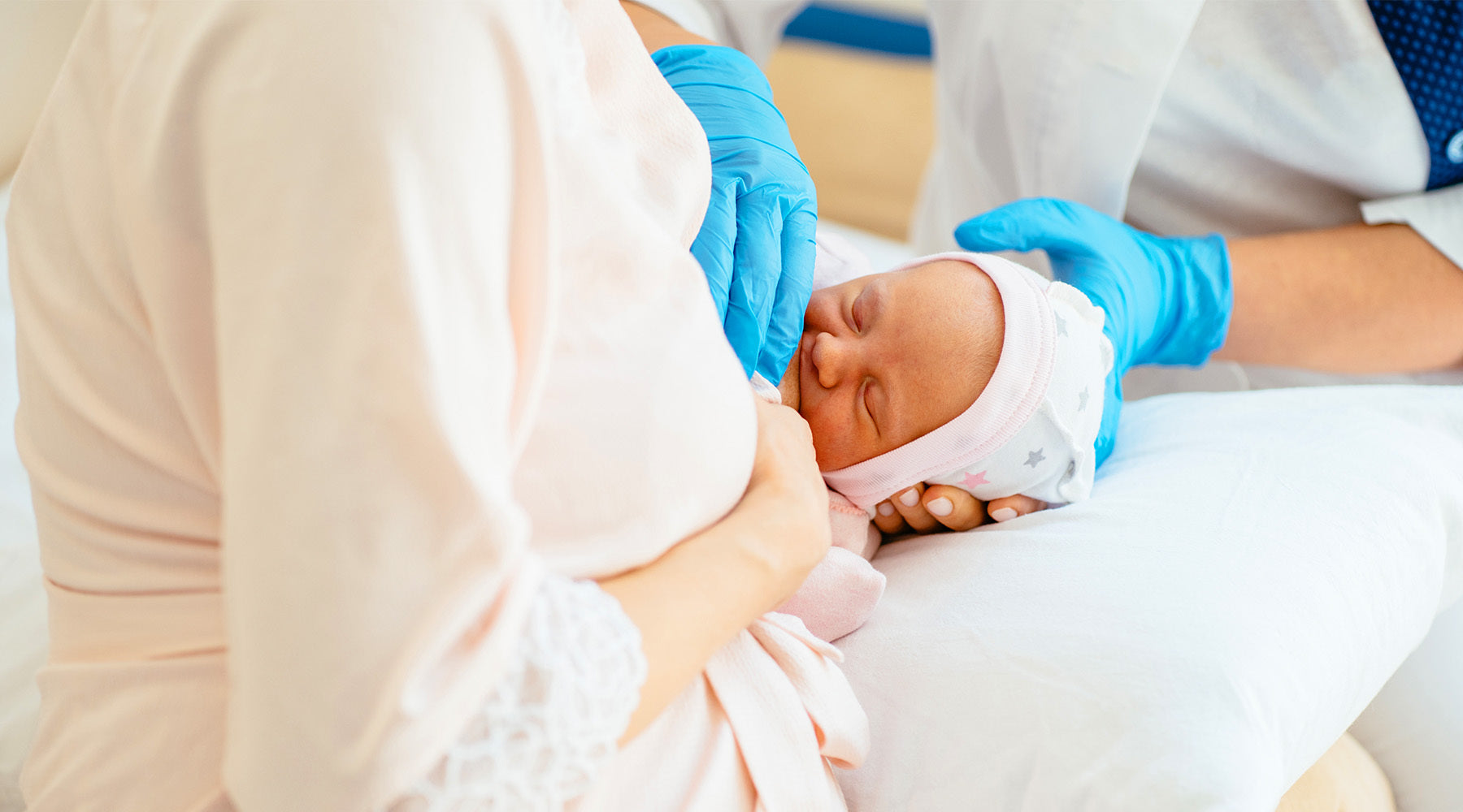 A newborn baby wearing a soft cap is gently held by a healthcare professional in blue gloves, while resting in the arms of a parent dressed in light pink clothing.