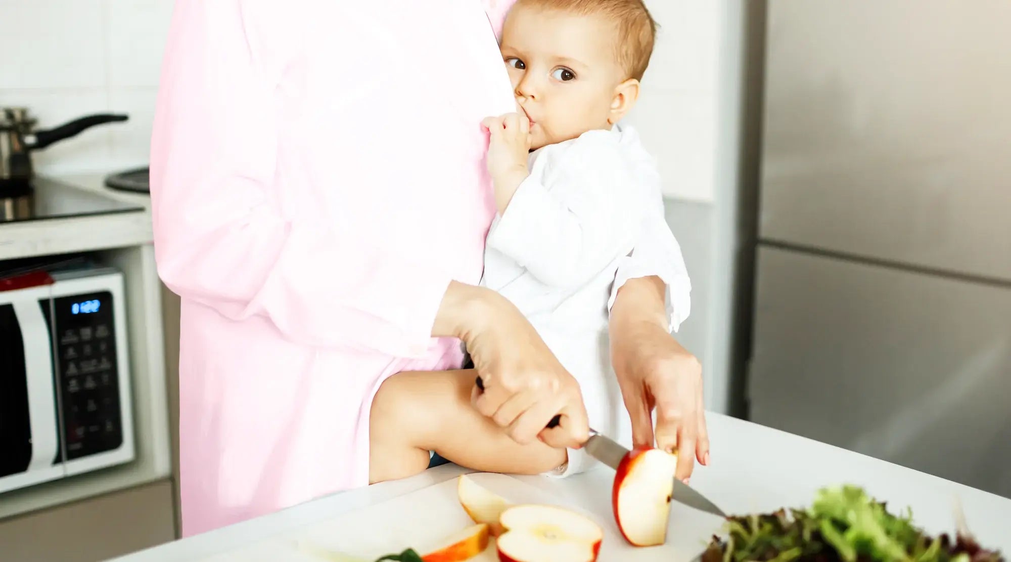 Mother holding baby while slicing fresh apples in the kitchen.