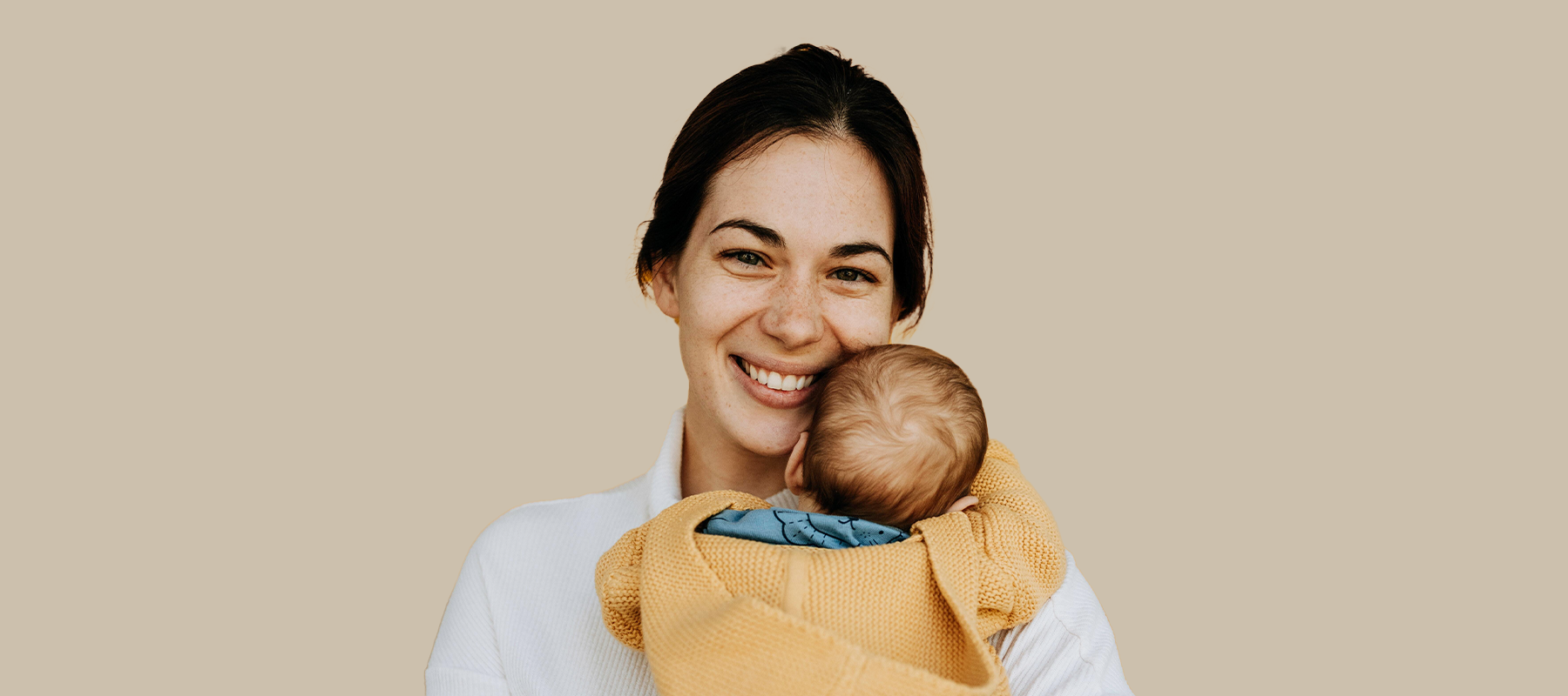 Smiling mother holding her baby wrapped in a yellow blanket against a neutral background.