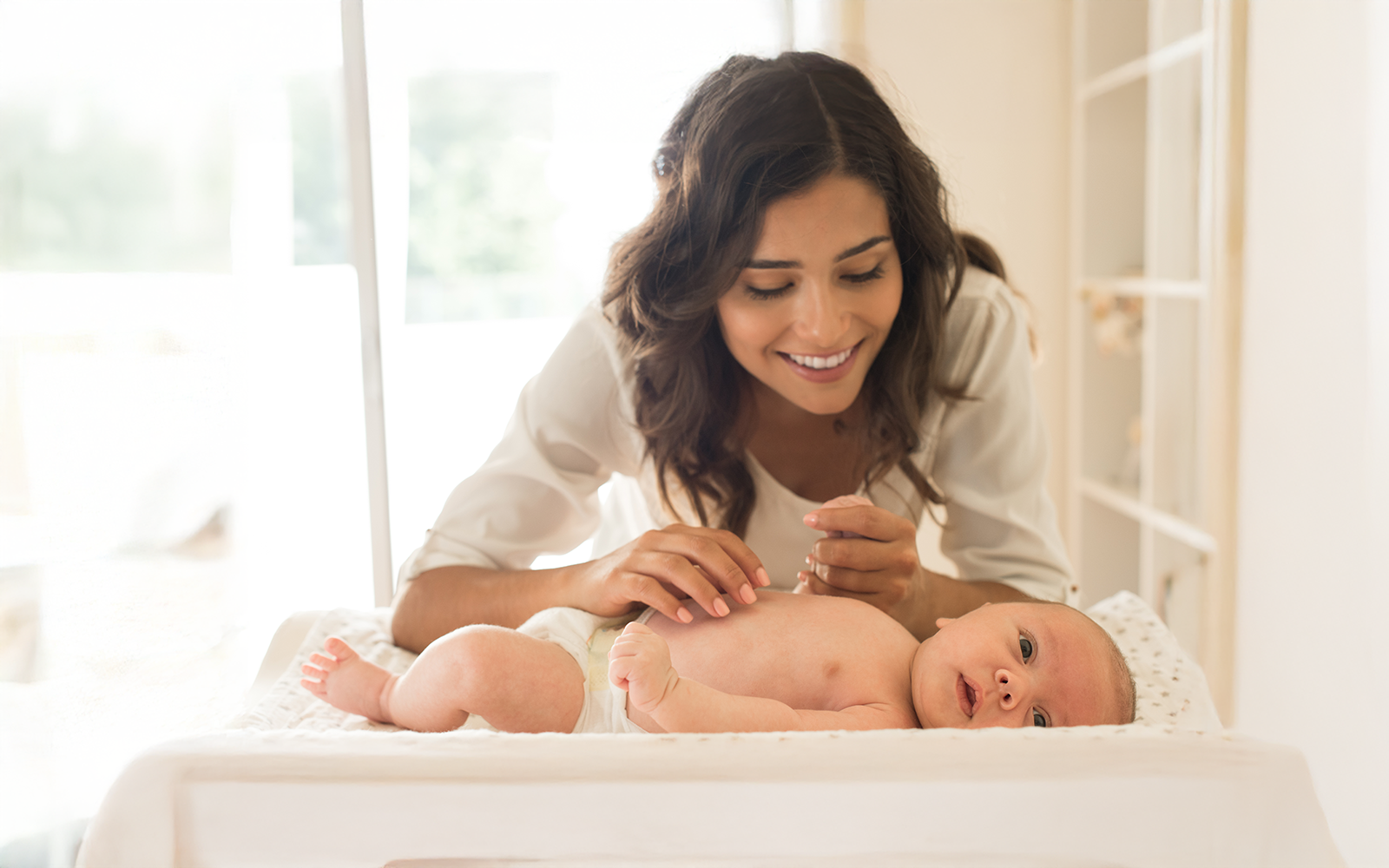 Smiling mother gently touching her baby lying on a changing table in a bright room.