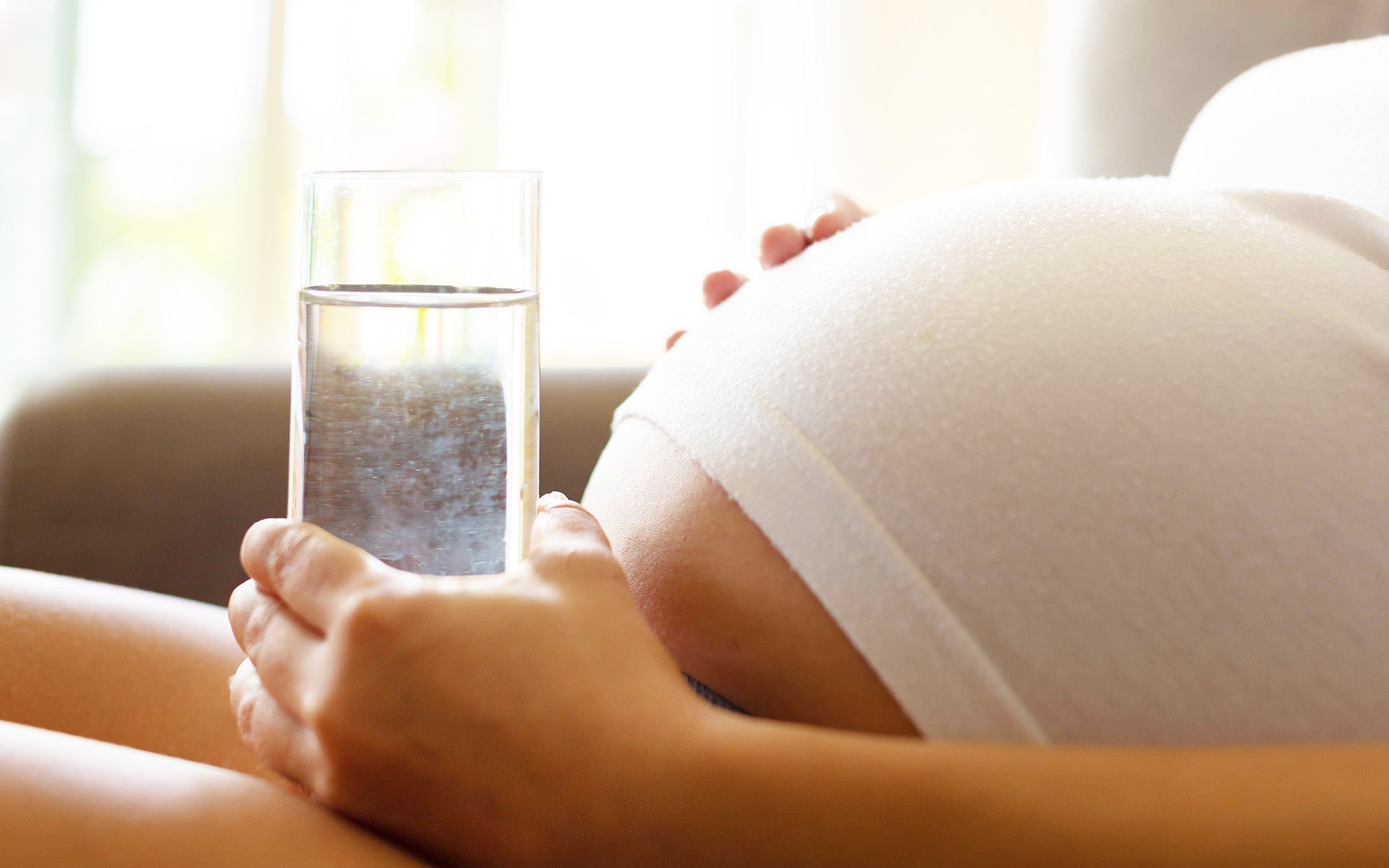 Pregnant woman holding a glass of water, close-up of baby bump in white clothing.