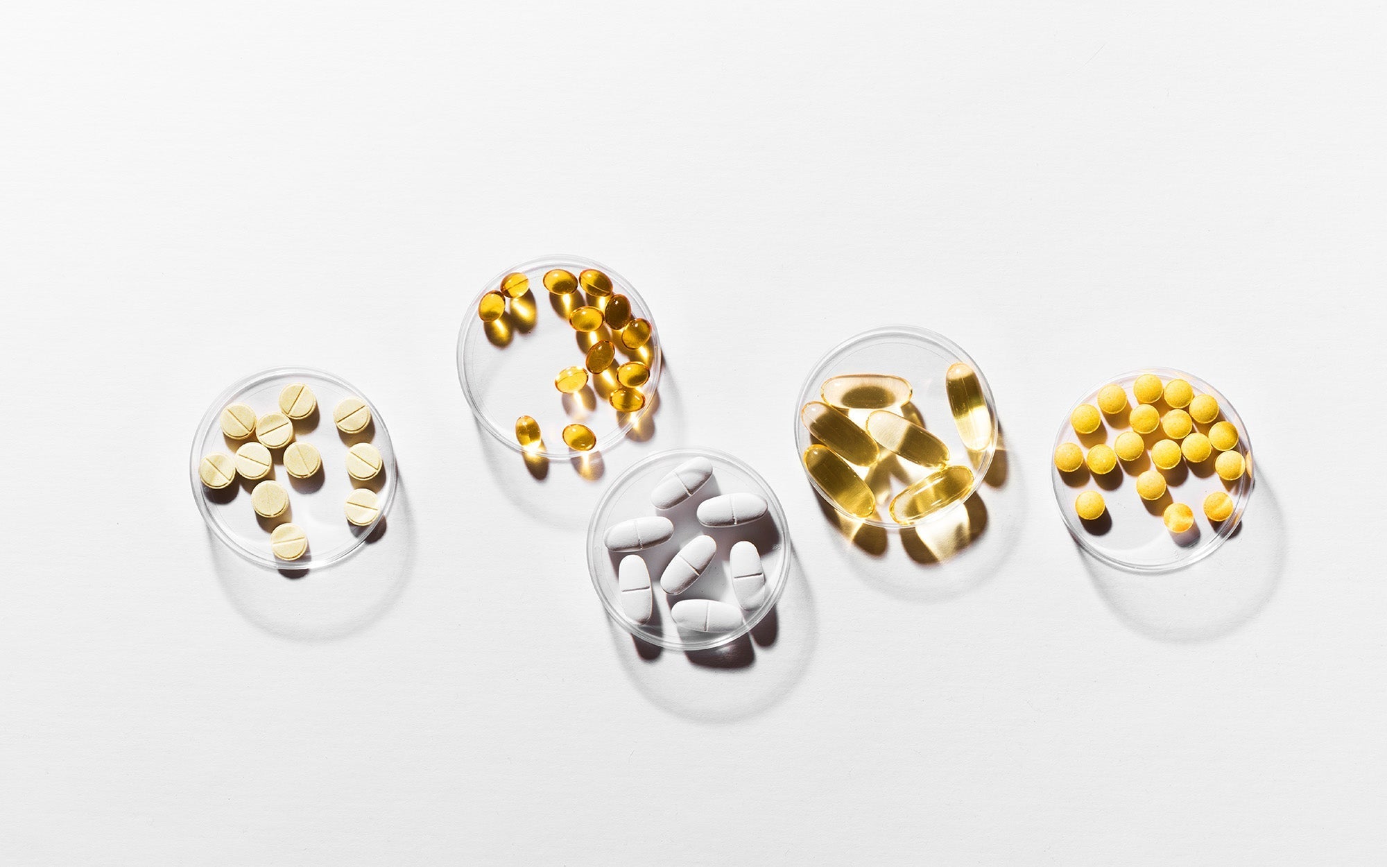 Assorted dietary supplement pills and capsules in small round dishes on a white background.