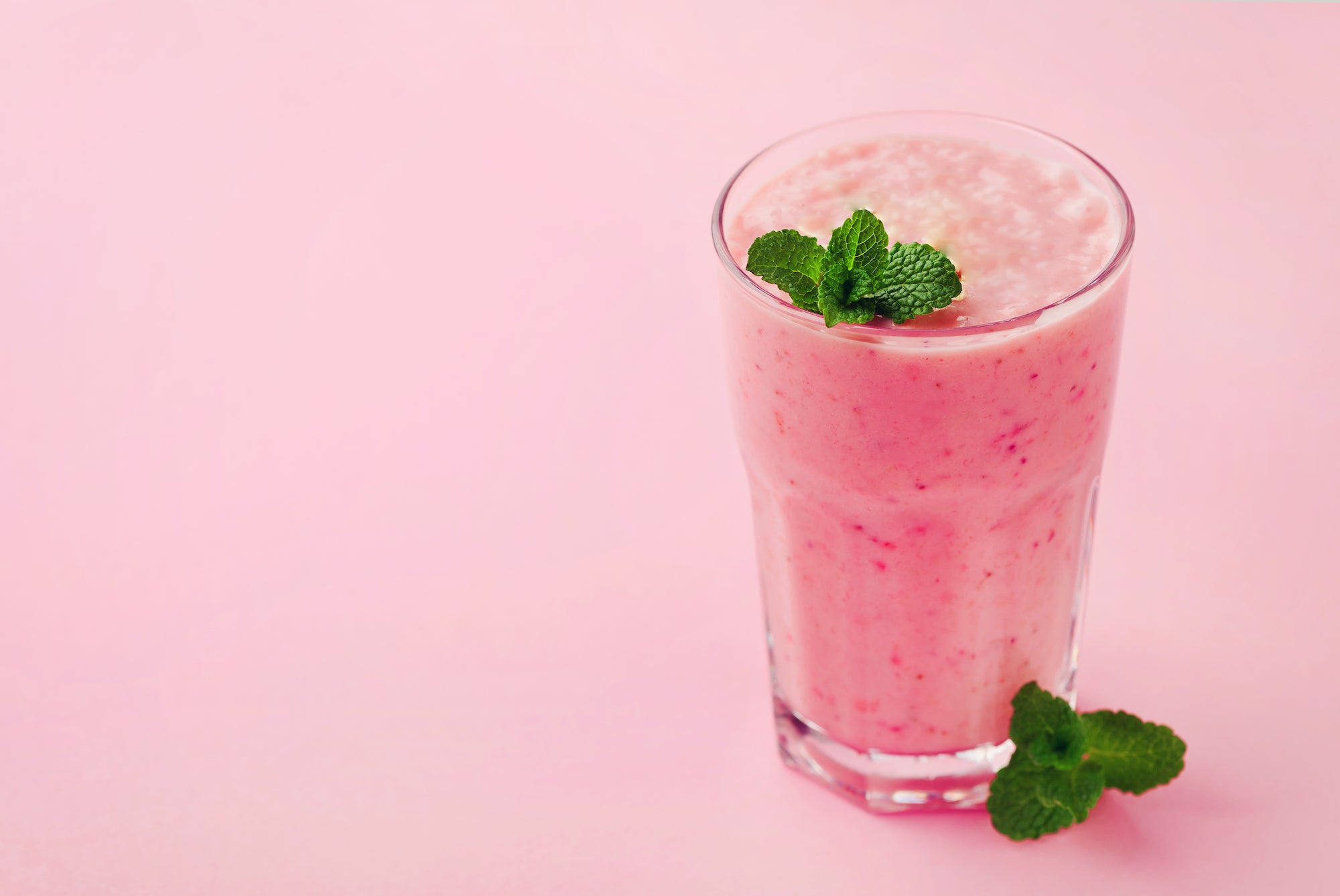 Glass of pink strawberry smoothie garnished with fresh mint leaves on a pastel pink background.