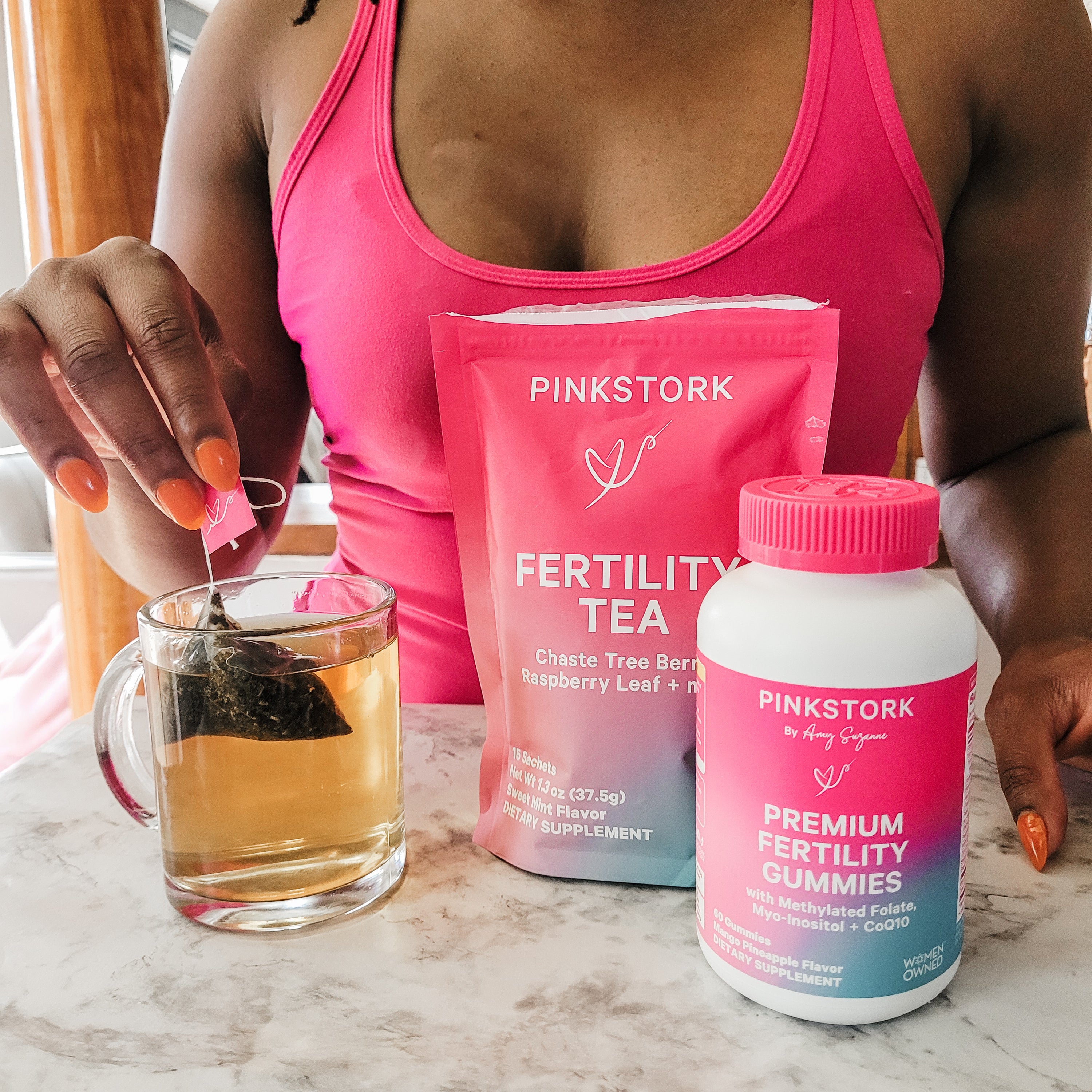 Person holding a glass of tea with Pinkstork fertility products on a marble surface