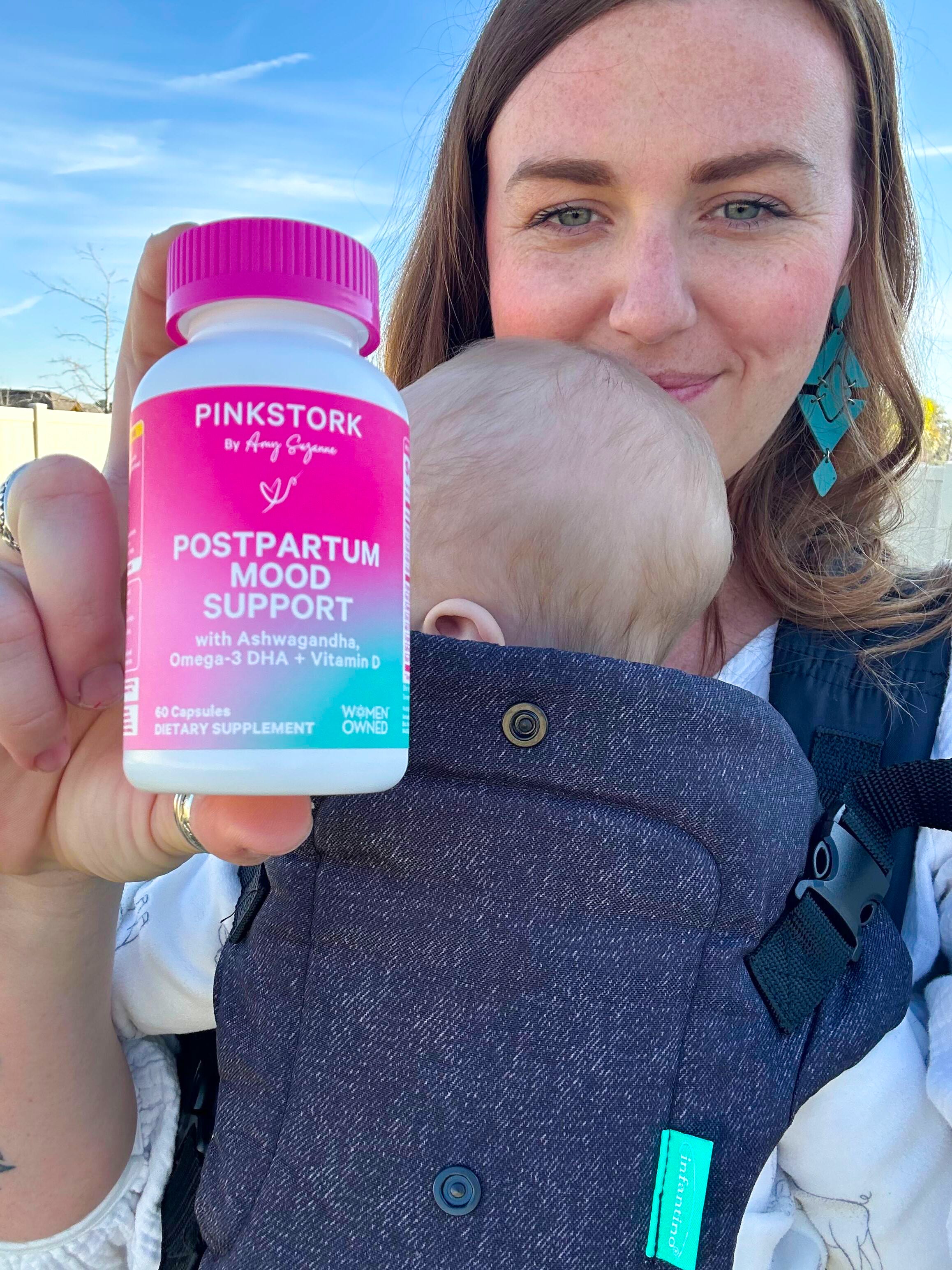 Woman holding a baby in a carrier and a Pink Stork Postpartum Mood Support supplement bottle.