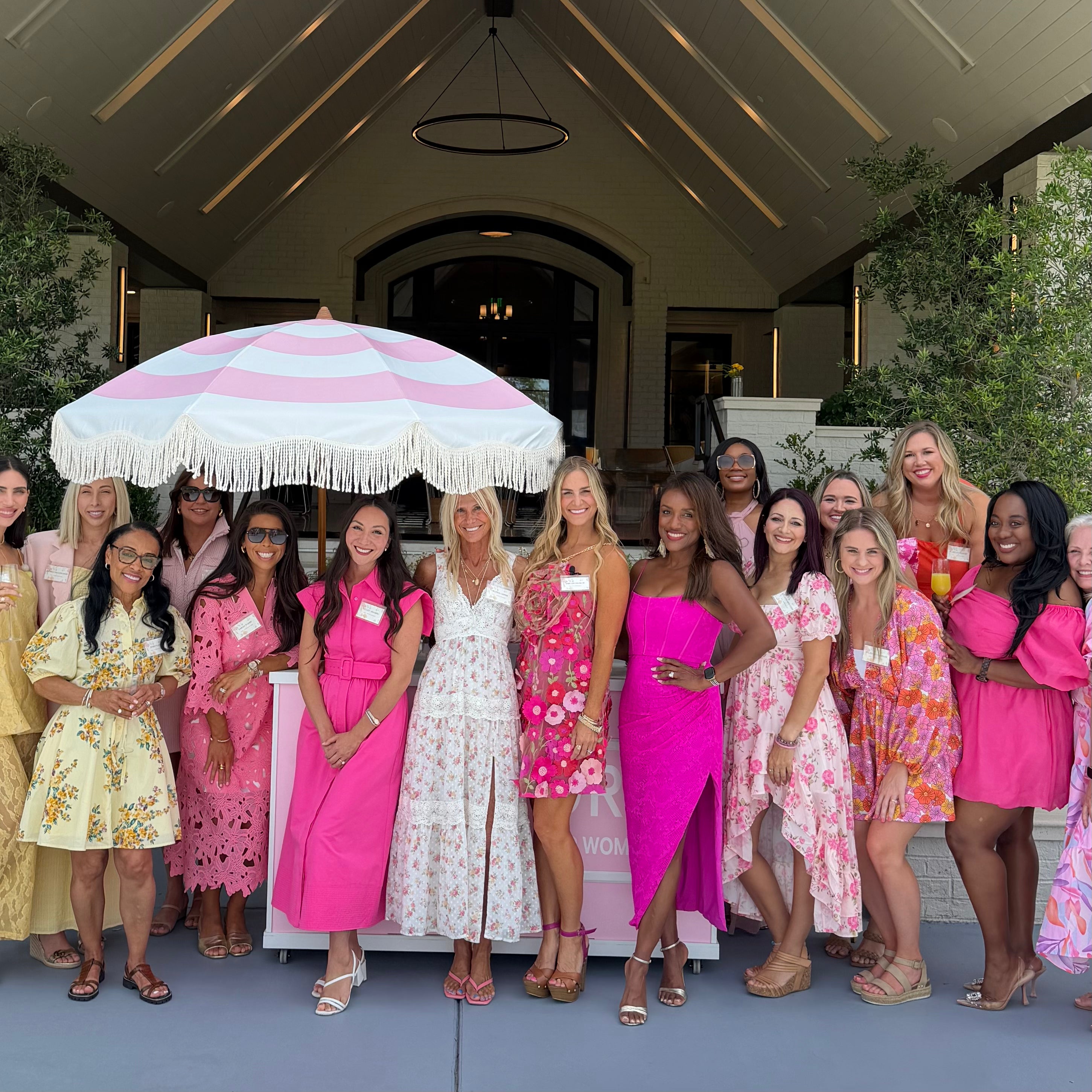 Women posing for a photo wearing pink, floral dresses surrounding a "Pink Stork" branded flower bar with a white and pink umbrella.