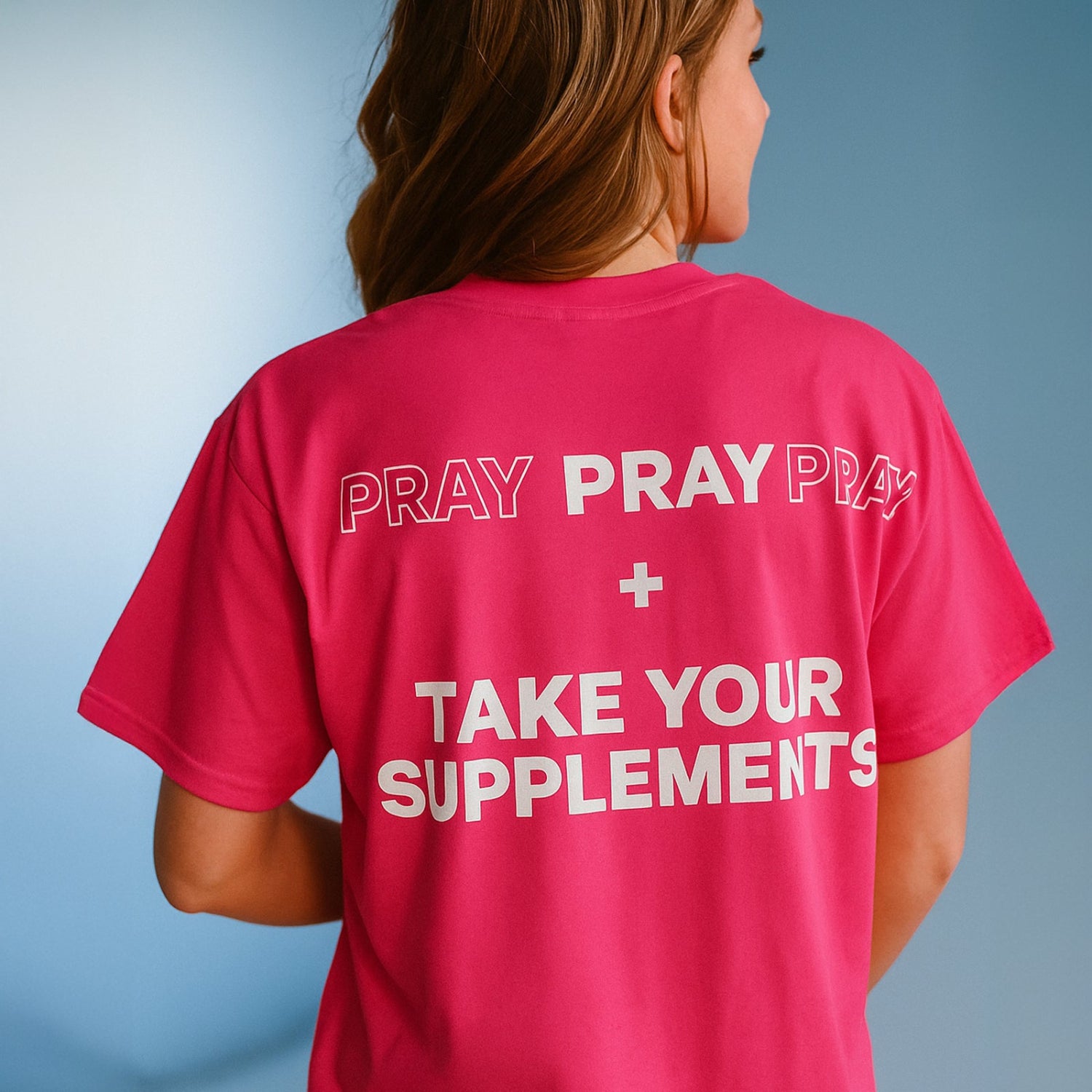 Person wearing a pink t-shirt with 'PRAY + TAKE YOUR SUPPLEMENTS' text on the back against a blue background