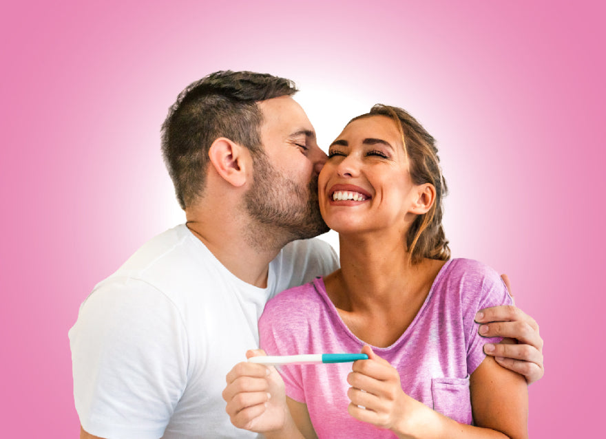 A white couple embrace. The woman is in a pink shirt and holding a pregnancy test. The man is in a white tshirt and is kissing her cheek. It's clear they have discovered they are pregnant, and elated.