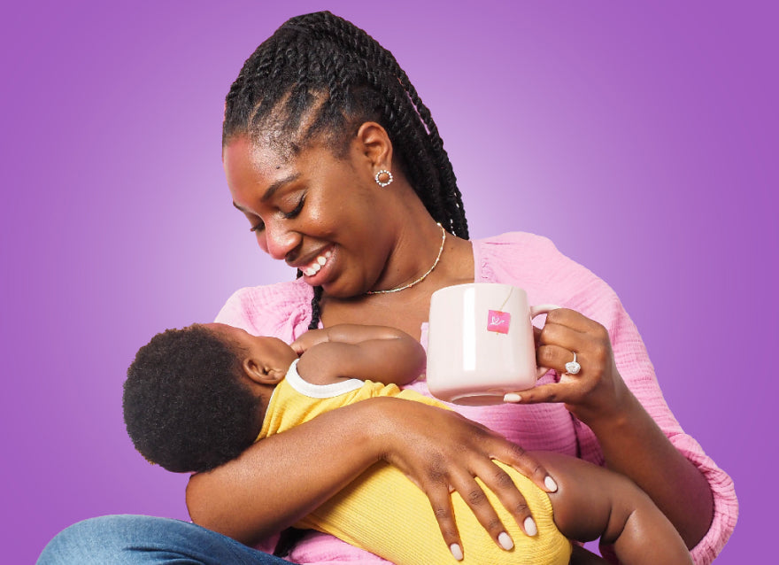 A black woman with braids pulled into a pony tail holds her baby, wearing a yellow onesie, to breast while she looks at them smiling. She holds a pink mug with a pink stork tea bag label hanging out the side.