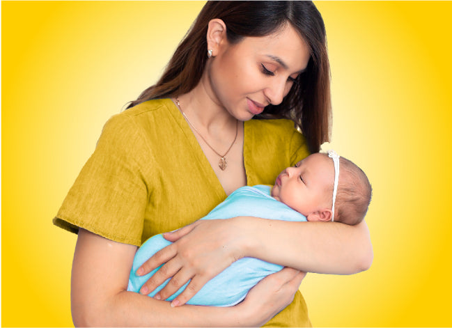 An olive skinned woman, wearing a yellowish top looks lovingly at her newborn who. wrapped in a blue blanket and wearing a white head band with a small yellow flower