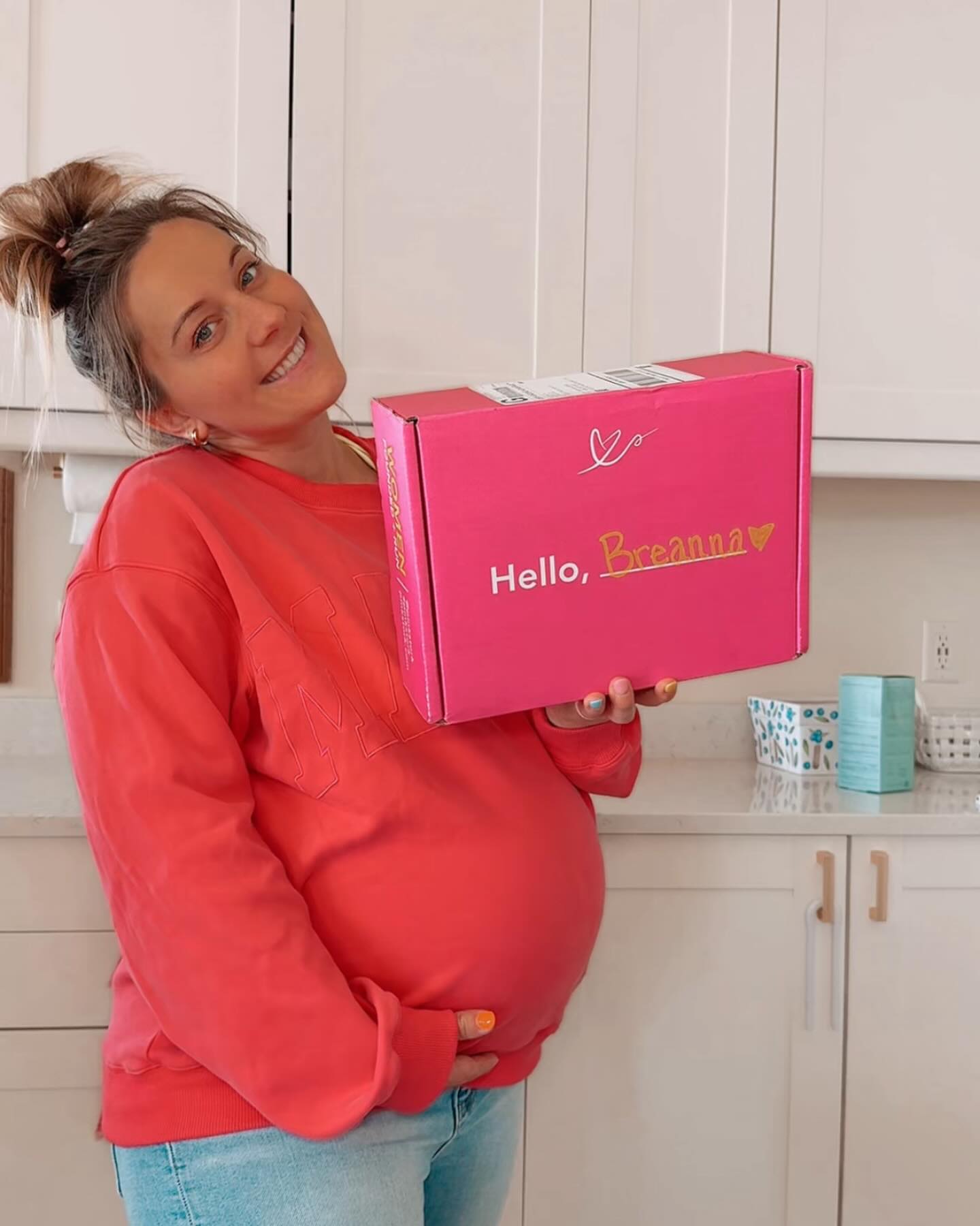 Pregnant woman holding a pink box with 'Hello, Baby!' text in a kitchen.