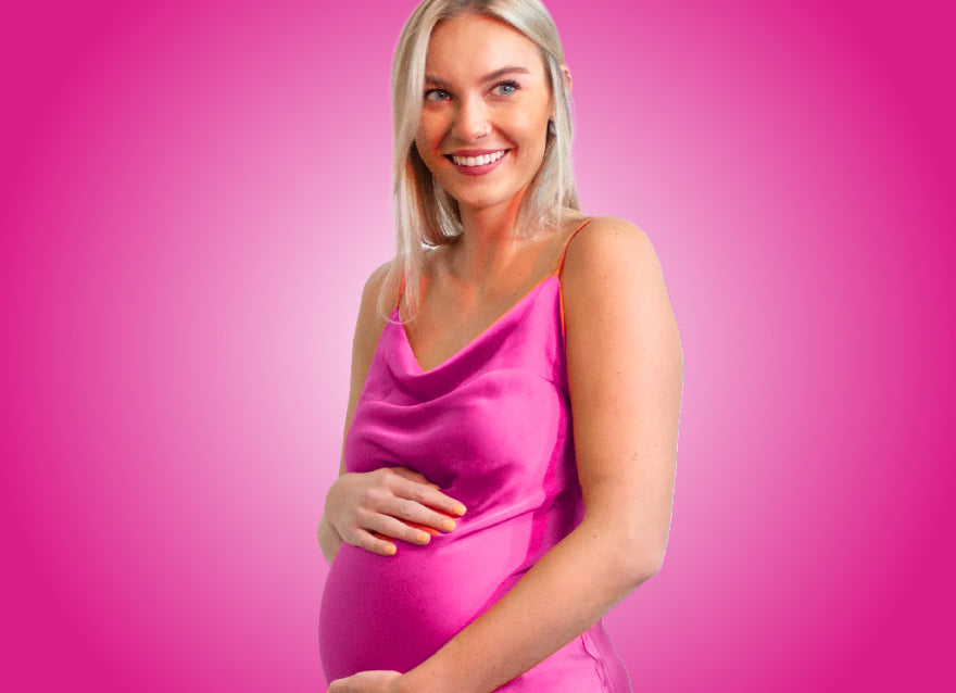 A happy looking white woman, with blonde hair, wearing a pink dress holds her pregnant belly.