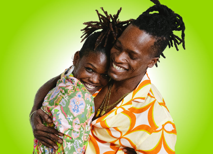 A black couple wearing colorful shirts, with locs pulled into ponytails hug and smile on a green background