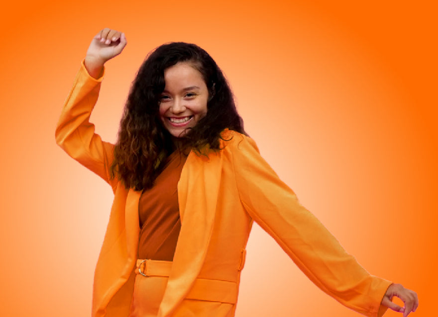 A latina woman with long brown hair, and wearing a casual orange suit strikes a dance-like pose, looking celebratory.